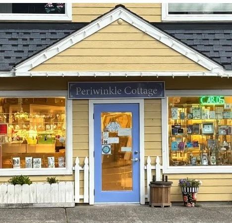 Yellow gift shop with blue door, large front windows displaying books and cards, and a sign reading 'Periwinkle Cottage' on the building.