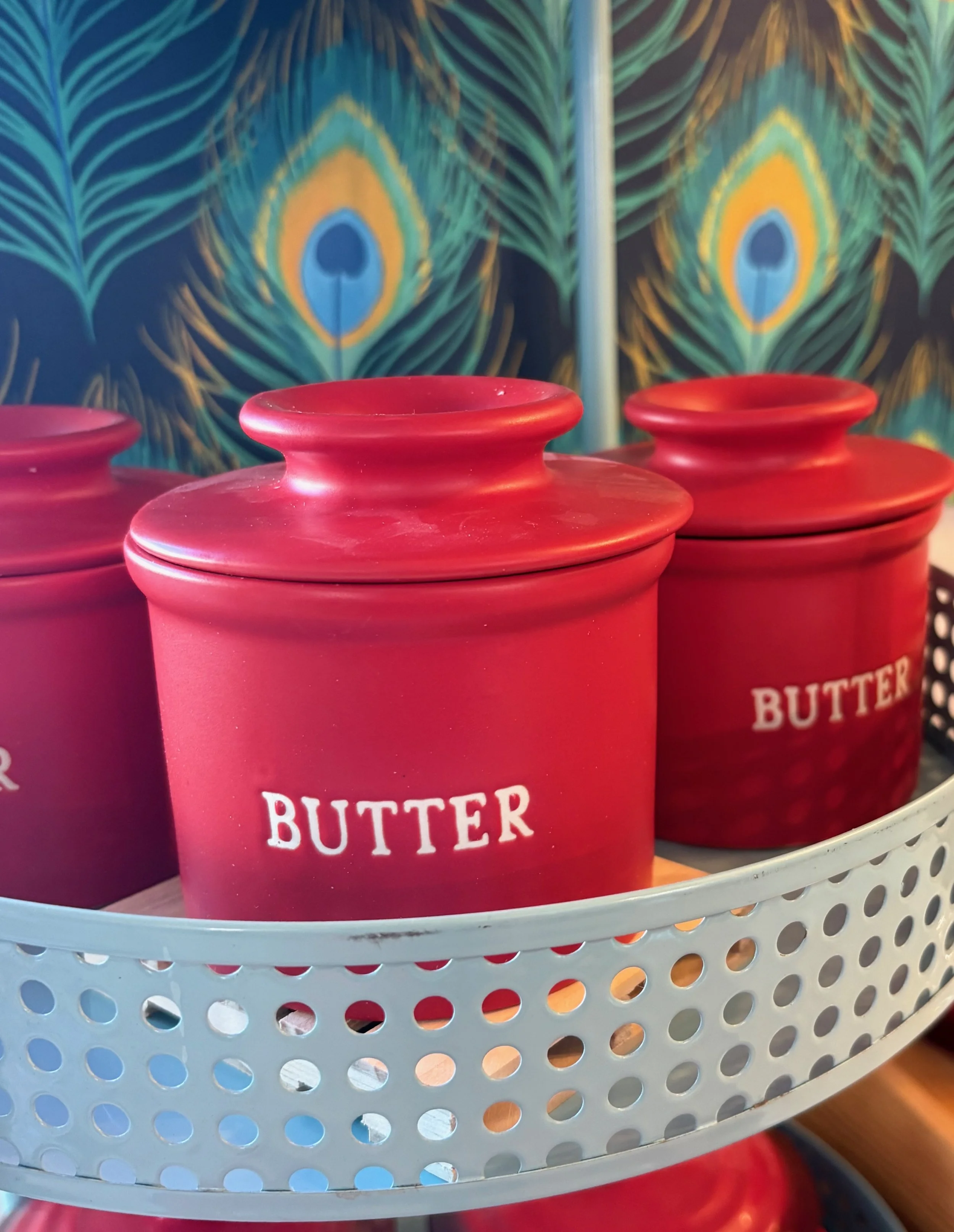 Red containers labeled 'butter' placed in a light blue circular basket with perforated holes, with peacock feather design wallpaper in the background.