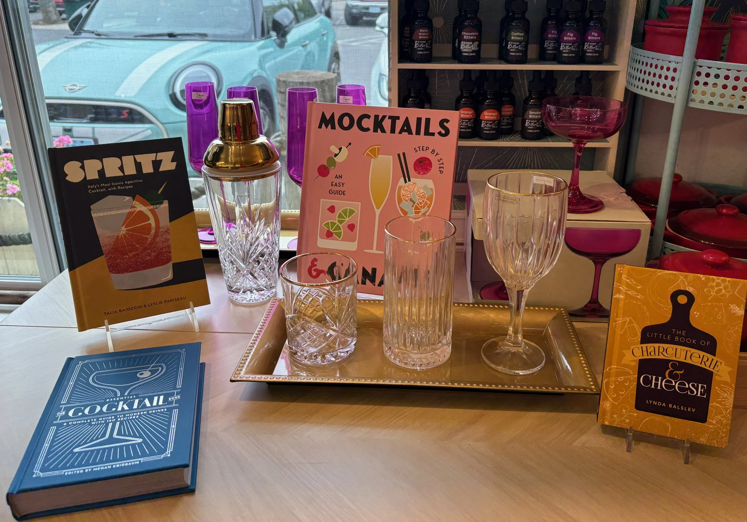 Display of cocktail recipe books and various glassware on a table inside a store, with a window showing parked cars outside.