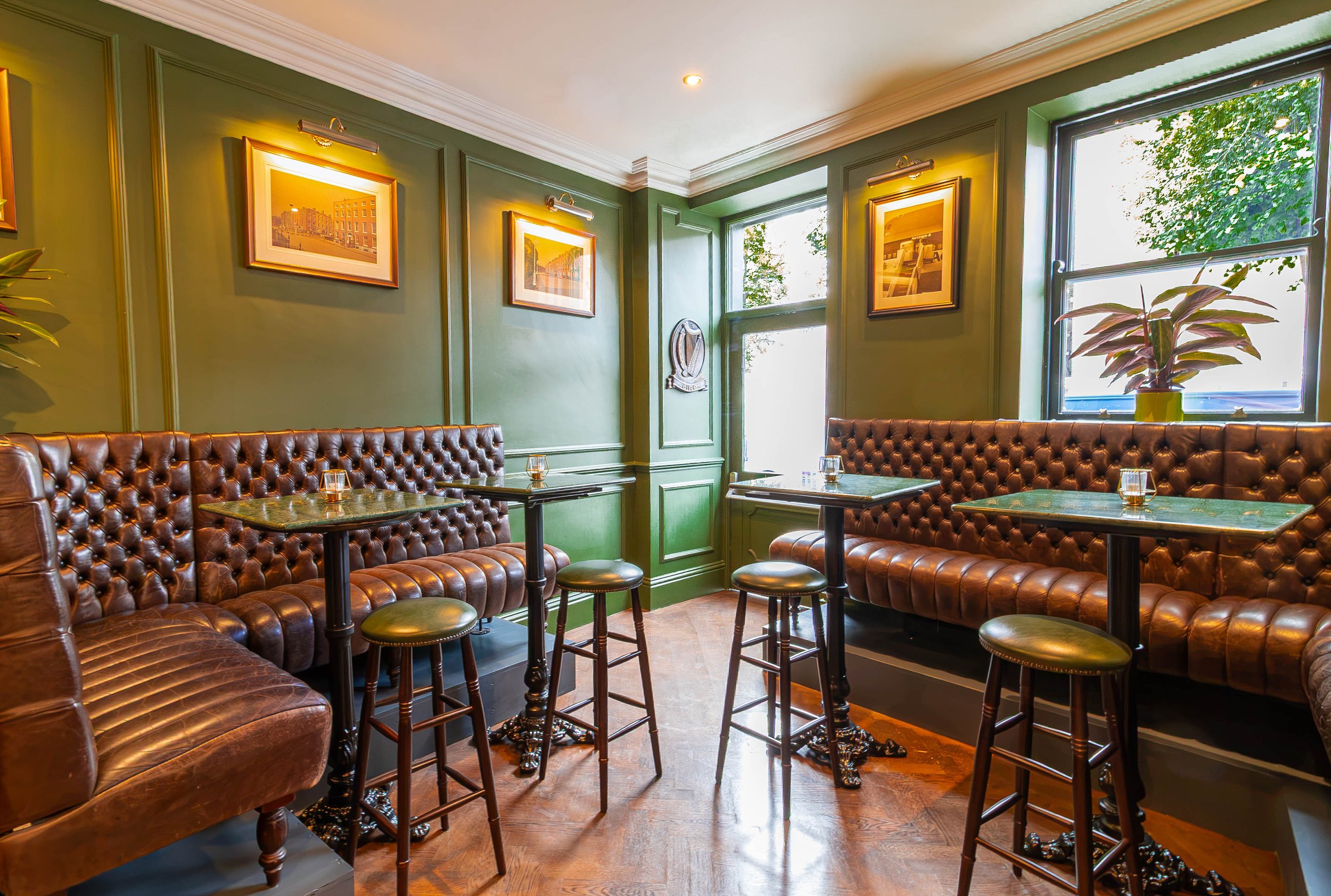 A cozy corner with green walls, framed pictures, and large windows allowing natural light. There are brown leather tufted sofas and small tables, along with a potted plant in the right corner. This image is in The Snug in Sally Mac's Pub on Baggot St