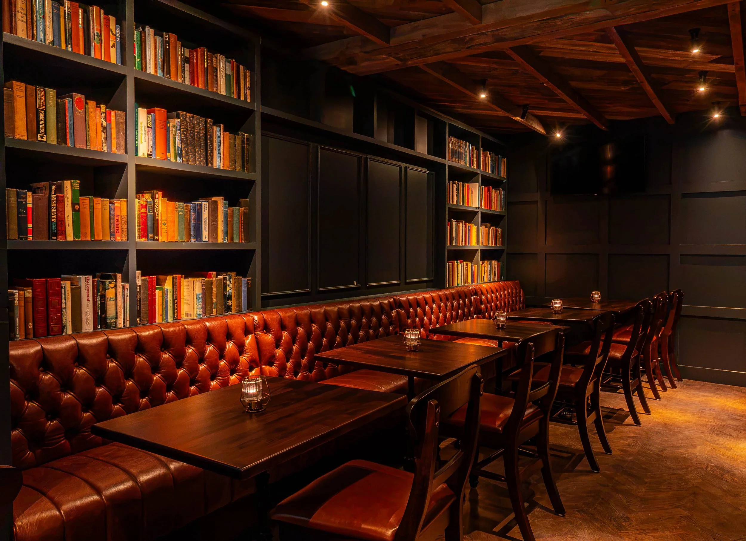 A cozy, dimly lit dining area with dark wooden tables, brown leather tufted banquette seating, and glasses on the tables. The background features black wall panels, a large wall-mounted TV, and tall bookshelves filled with colorful books, with a wood-paneled ceiling and small ceiling spotlights.