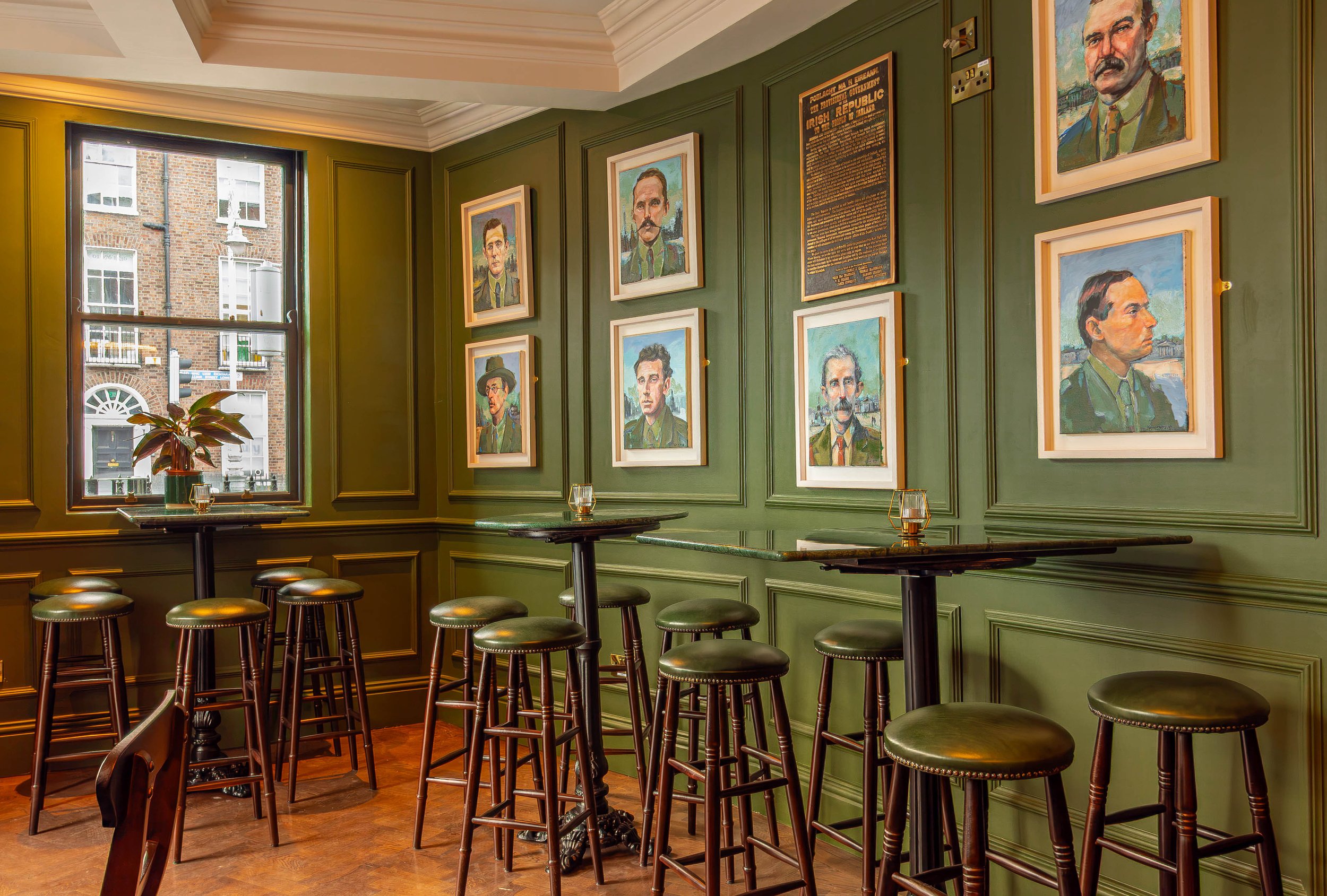 Interior of a cozy pub with green paneled walls decorated with framed paintings of historical Irish figures, a window showing brick buildings in Dublin, and high wooden stools around tall tables. Taken in Sally Mac's Pub & Kitchen on Lower Baggot St.