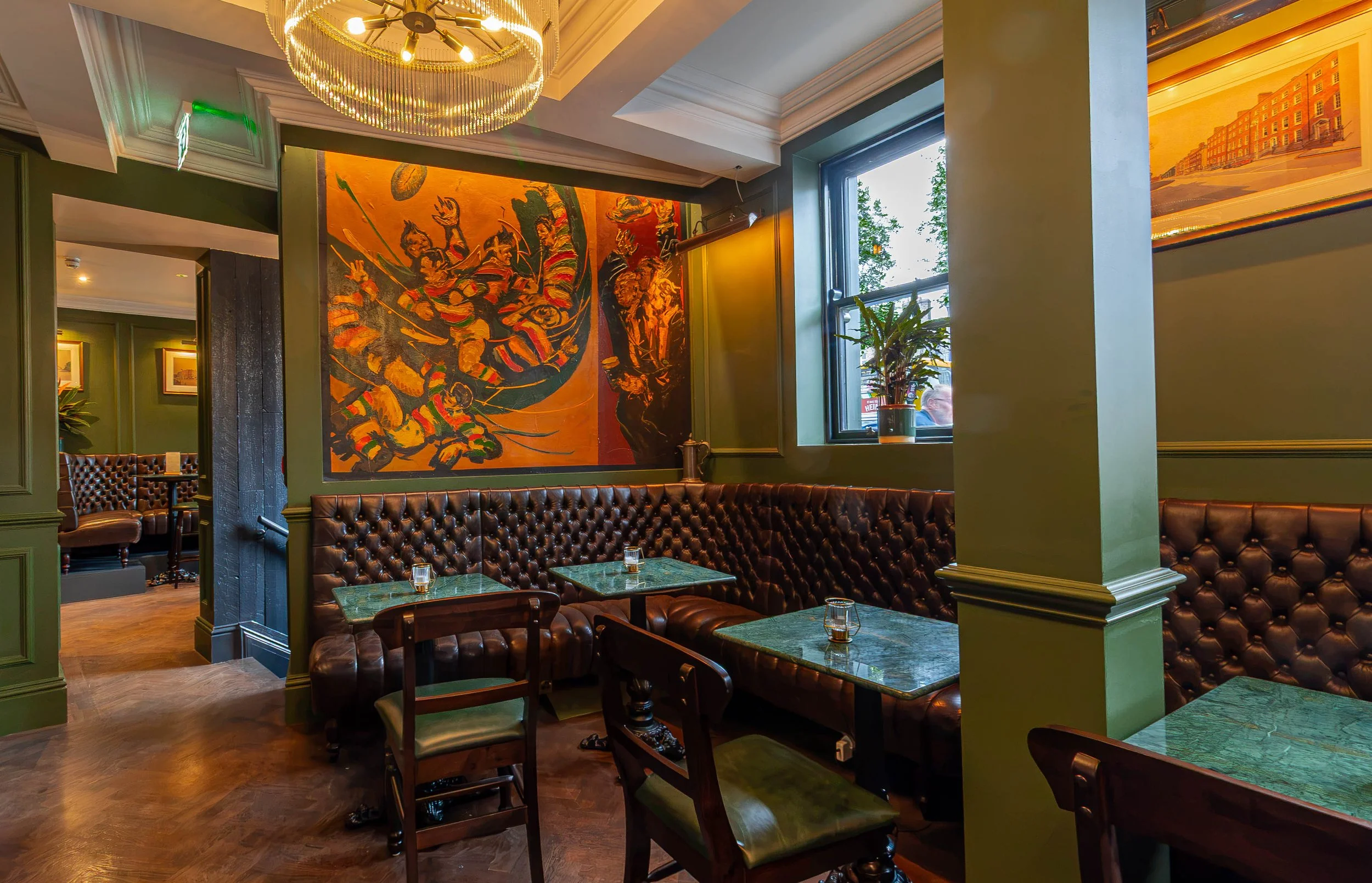 Interior of Sally Mac's Pub in Dublin with dark green walls, brown leather tufted booths, and green marble tables. There is a large, colorful mural painted by Walsh, and a window letting in natural light. The ceiling has a gold chandelier.