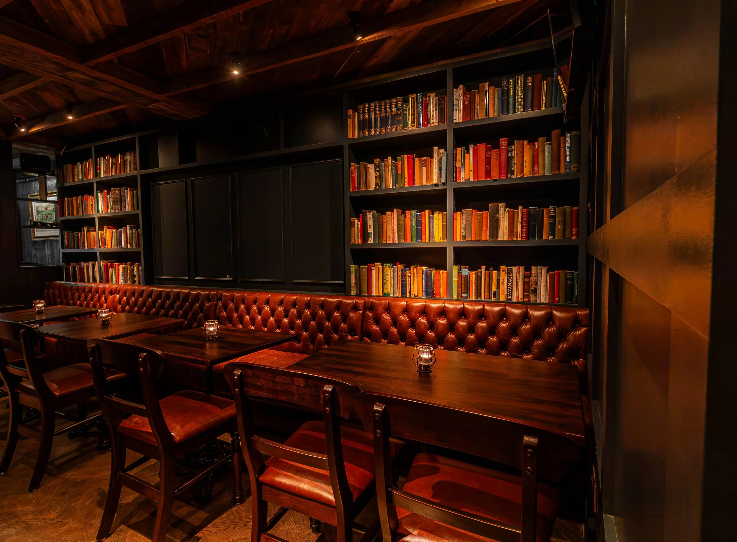 Sally's Writer's Room Lounge has dark walls and a wooden ceiling, filled with bookshelves and warm lighting. There is a long, brown leather tufted sofa along the wall with several small wooden tables and chairs, creating a warm atmosphere.