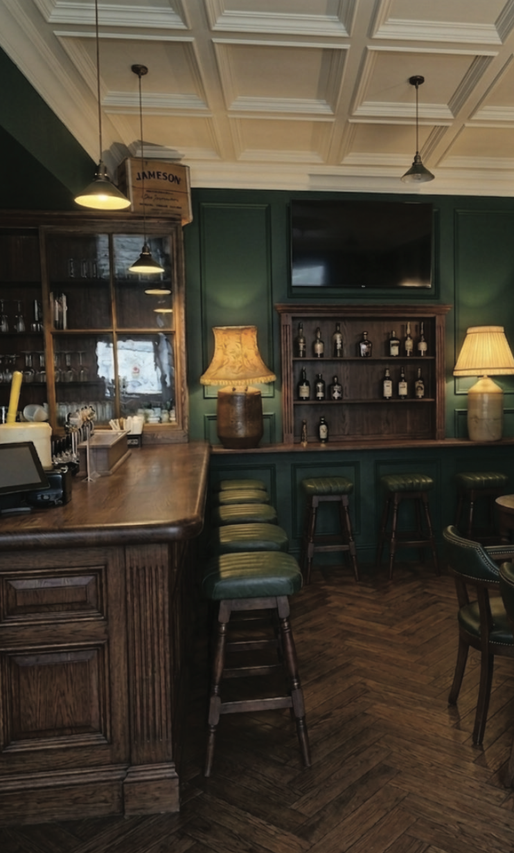 Interior of a bar with wooden furniture, green walls, and a lamp on the counter. There are bar stools with green cushions, a TV above a shelf of bottles, and warm lighting.