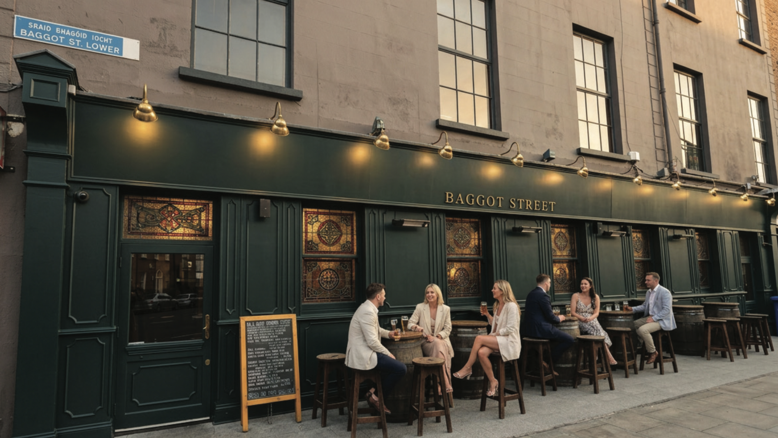 People enjoying drinks outside a pub named Baggot Street with a green exterior, stained glass windows, and hanging lights, on Baggot Street Lower in Dublin.