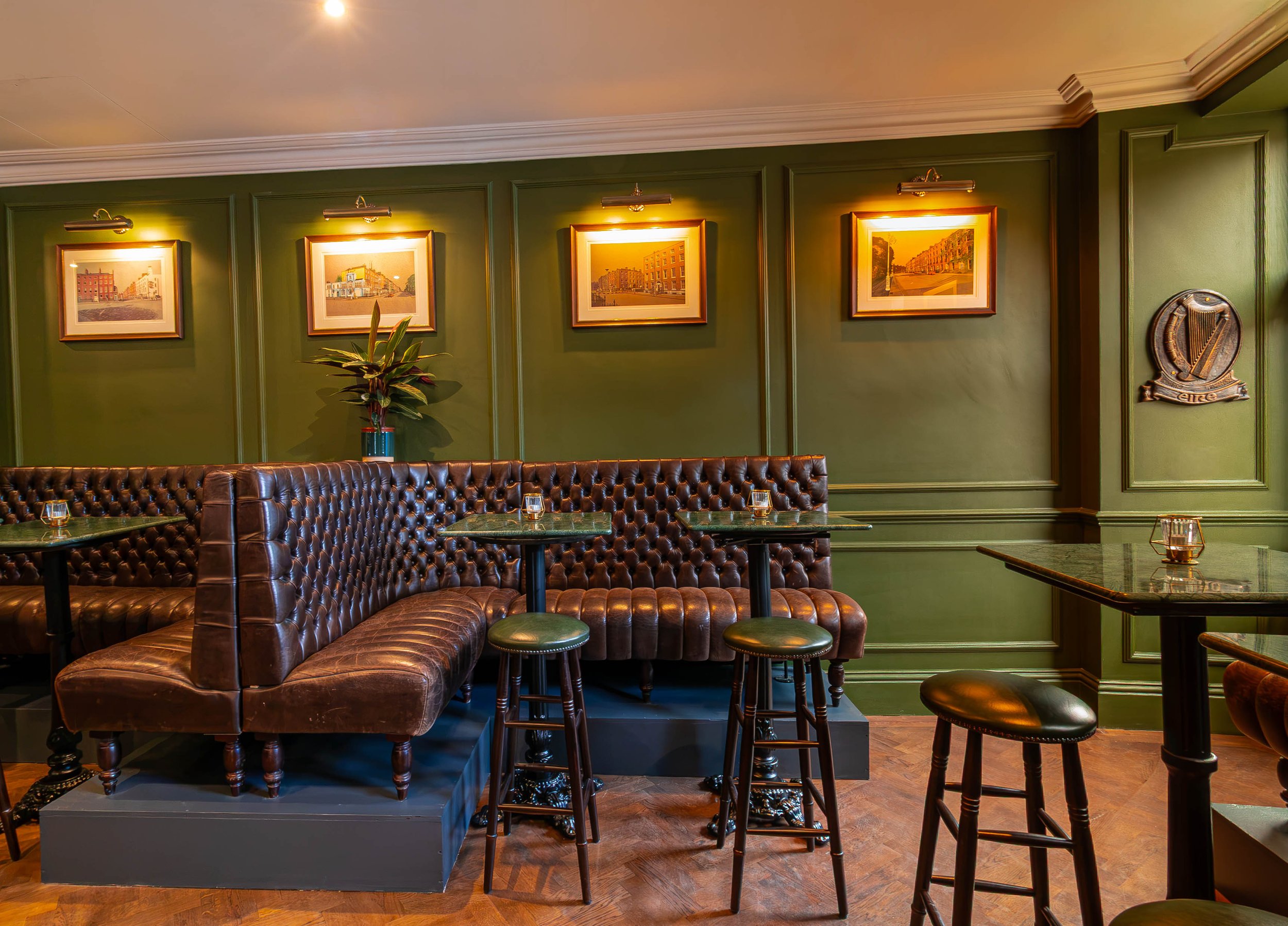 Interior of Sally Mac's Pub & Kitchen on Lower Baggot Street Dublin 2. Green walls, framed artwork of Dublin City Buildings illuminated by picture lights, brown leather tufted booths, and green marble tables with tealight candles.