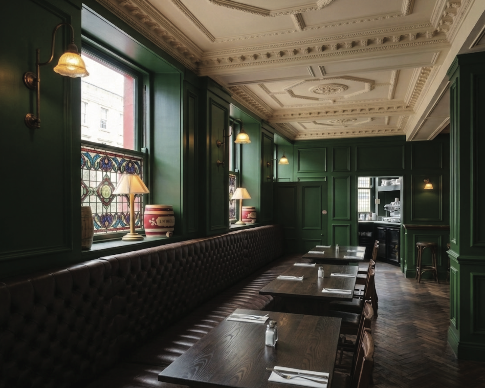 A cozy restaurant interior with dark green paneled walls, a stained glass window, and a long tufted leather banquette along the wall. Wooden tables are set with napkins, silverware, and salt shakers. Small lamps on the window sill and wall sconces pr