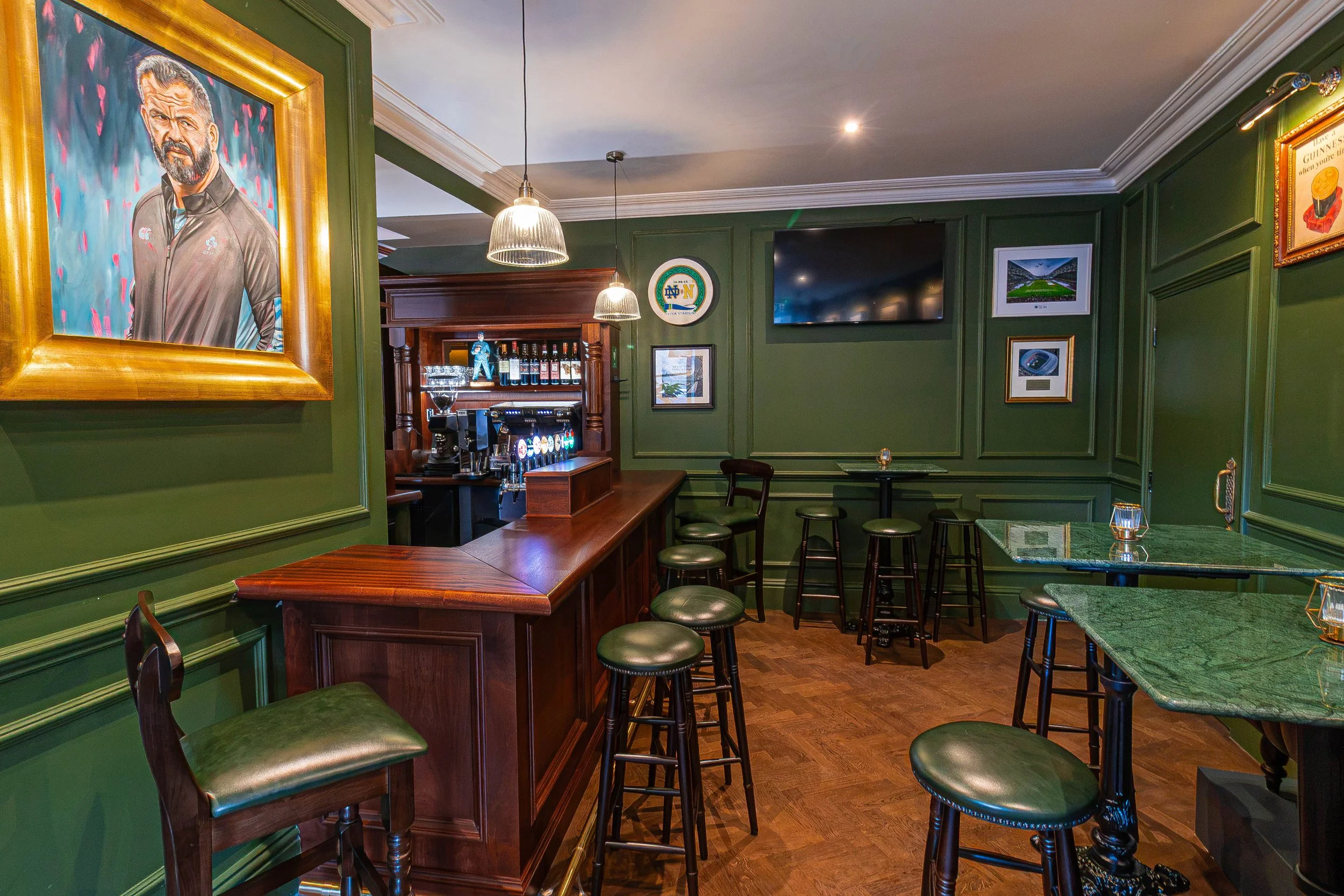 Interior of The Snug a section of Sally Mac's Pub & Kitchen on Lower Baggot St, Dublin 2. With a wooden bar, green leather stools, framed pictures, and a large portrait of Irish Rugby Player Andy Farrell, and a television is mounted on the wall.