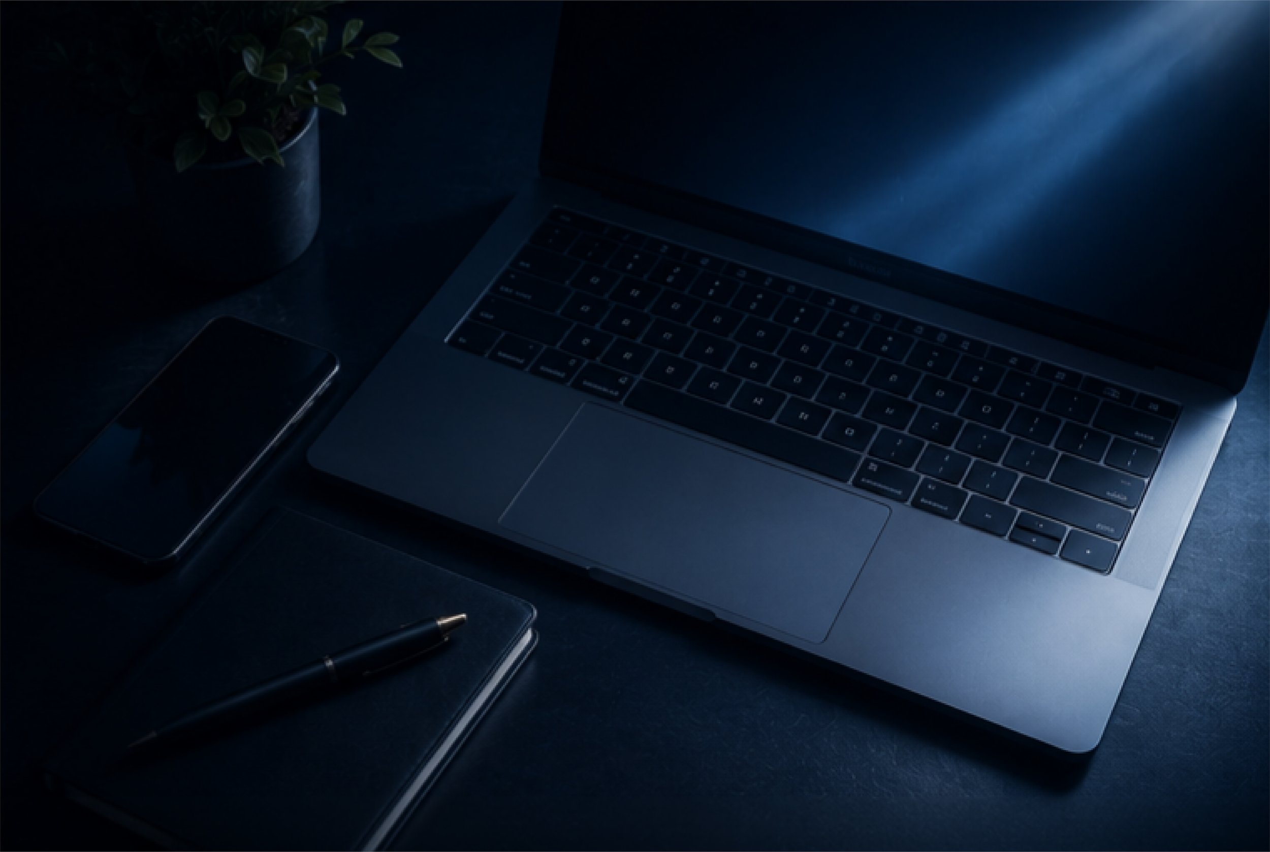 A dark workspace featuring an open laptop with a backlit keyboard, a closed smartphone, a pen, a small notebook, and a potted plant on a dark surface.