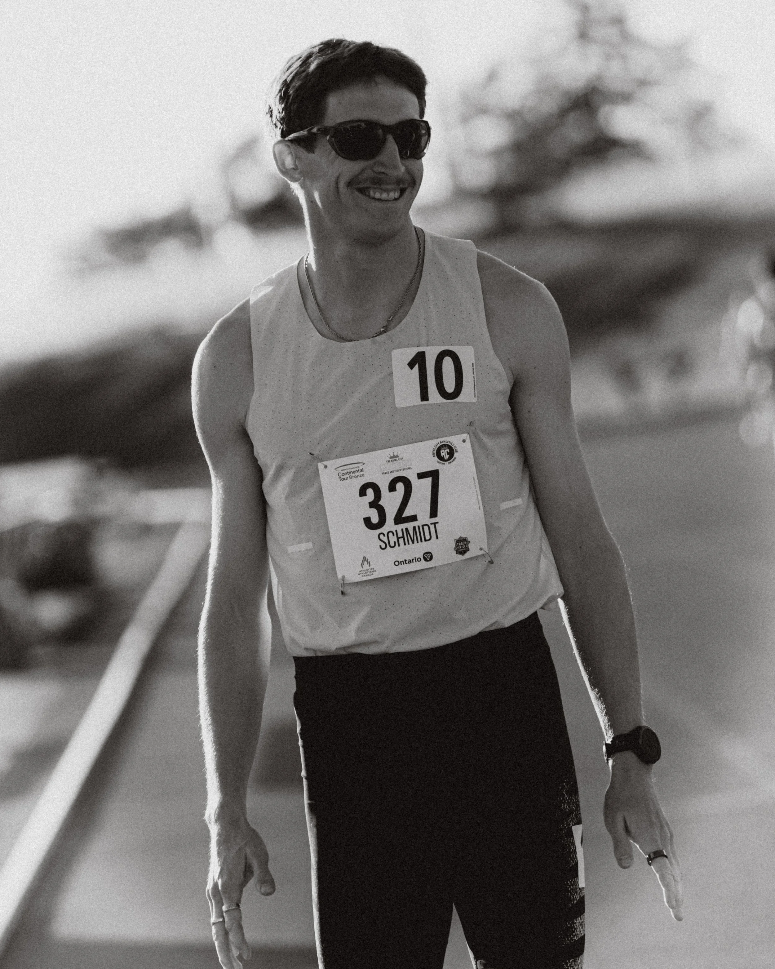 A smiling man wearing sunglasses, a sleeveless athletic shirt with a bib number 327 labeled Schmidt, standing outdoors with trees in the background.