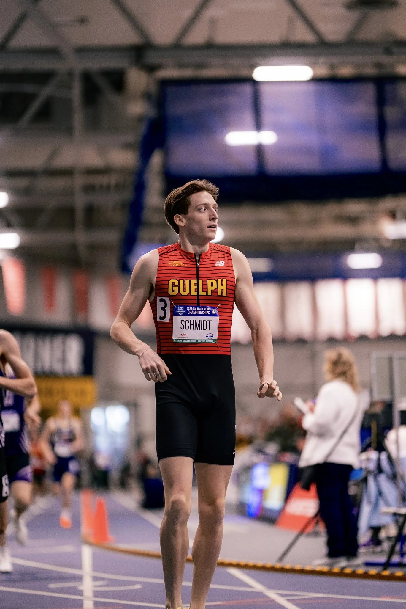 A male athlete wearing a red and black Guelph jersey with bib number 3, standing on an indoor track at a competitive event with other athletes and spectators in the background.