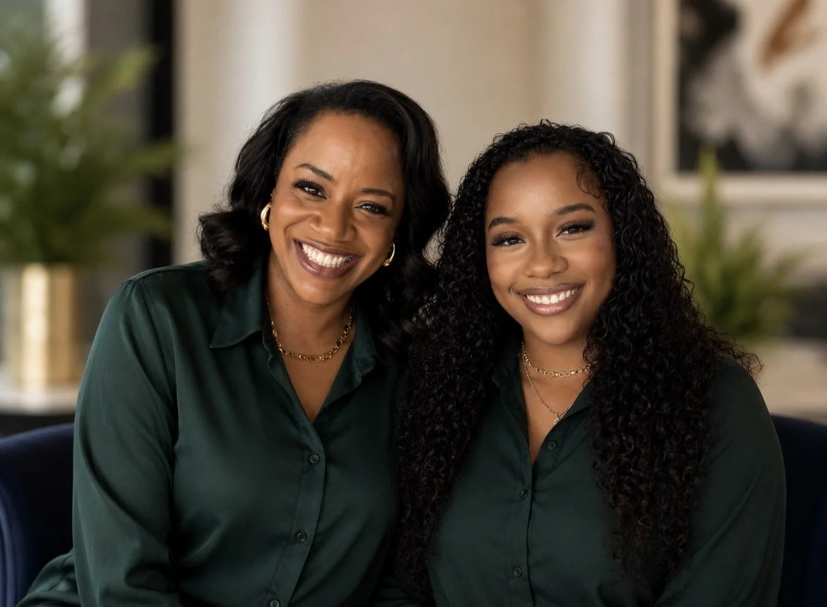 Two women smiling and sitting together indoors, both wearing dark green blouses and gold jewelry.