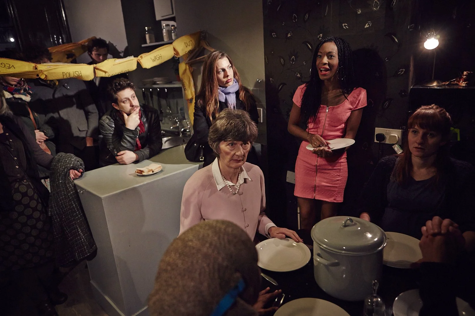 Group of people gathered in a dimly lit kitchen or dining area, with a woman in a pink dress holding a plate, speaking or explaining something, while others listen attentively. There are dishes, a pot, and a loaf of bread on the counter.