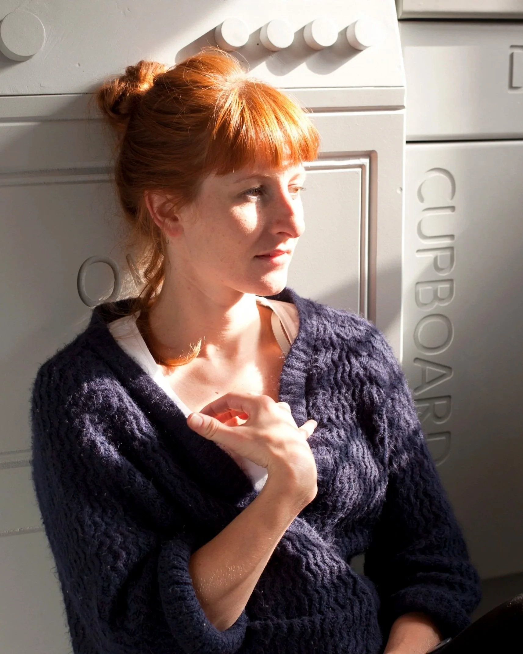 A woman with red hair styled in a bun and bangs, wearing a dark purple knitted sweater, is looking to her left in sunlight. She is in front of a white appliance with the brand name 'CUPBOARD' visible.