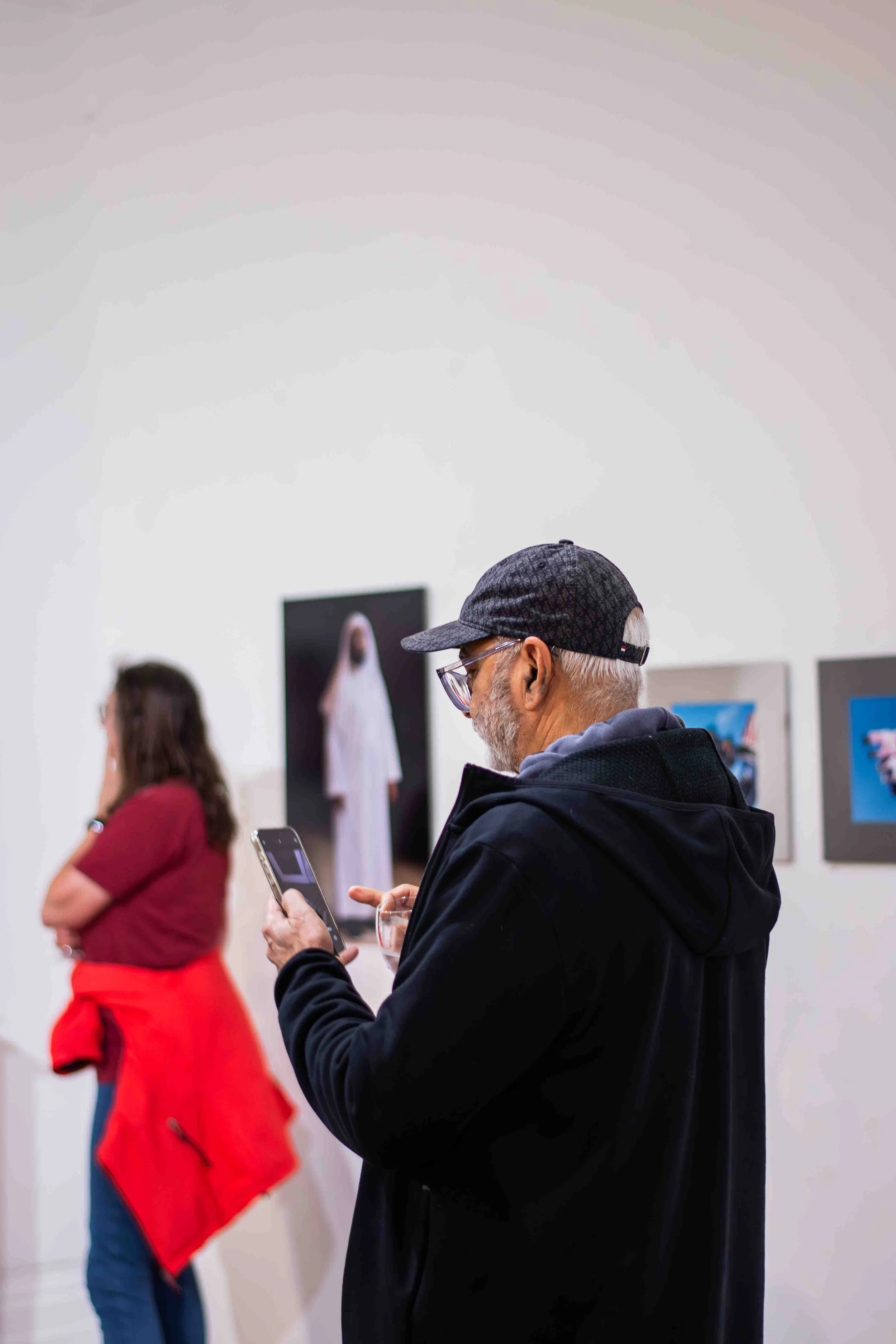 An older man wearing glasses, a dark cap, and a black jacket is looking at his phone in an art gallery. Behind him, a woman with long brown hair, a red top, and blue jeans is standing with her arms crossed, observing artwork. Several framed pieces ar