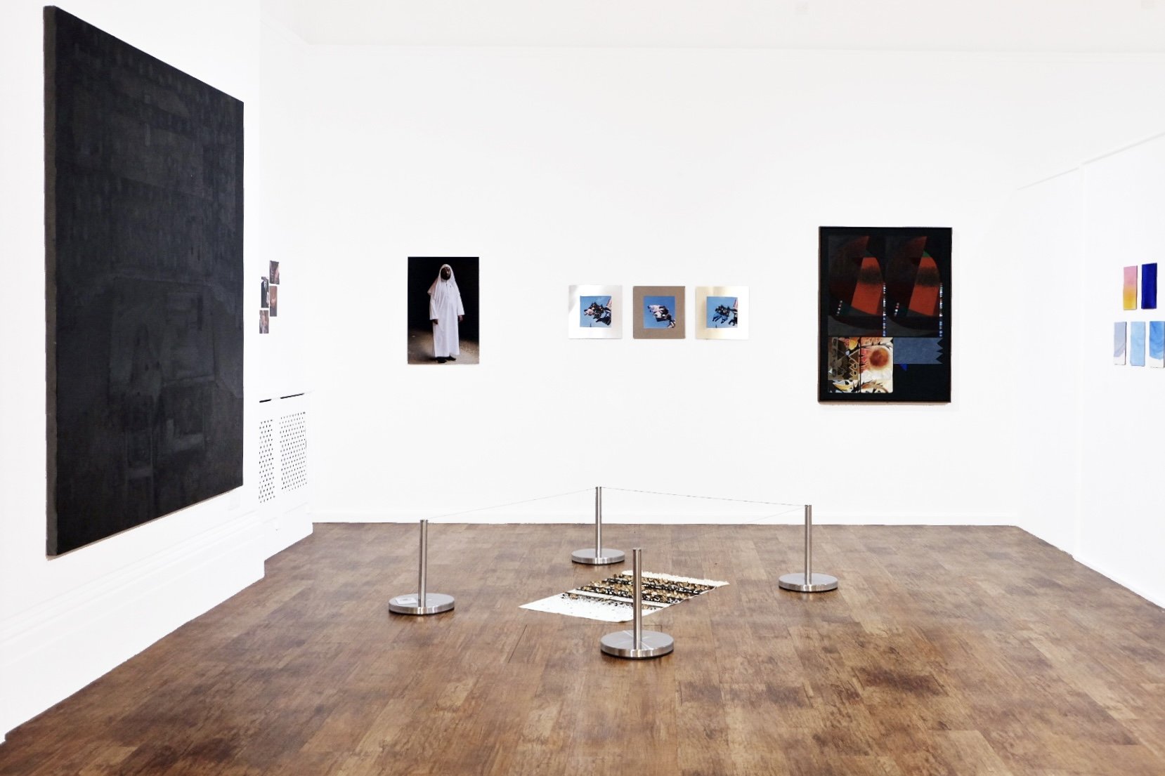 An art gallery with white walls displaying various paintings and photographs, wooden floor, and a small floor tapestry in the center surrounded by four stanchions.
