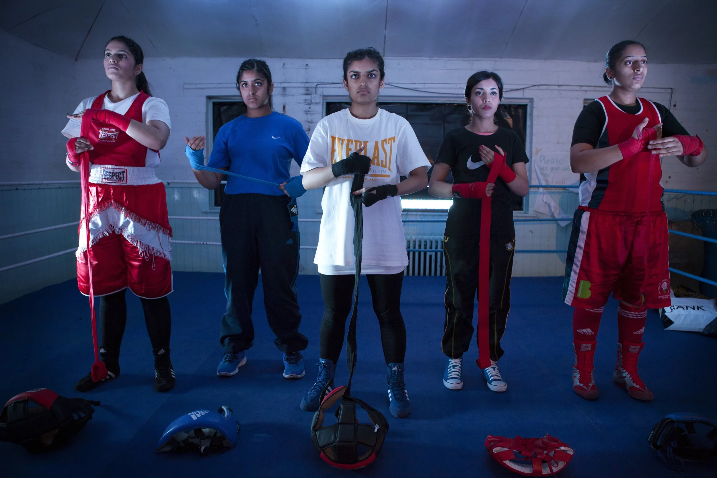 Five young female boxers standing in a boxing gym, dressed in training gear, with boxing gloves and headgear on the floor in front of them.