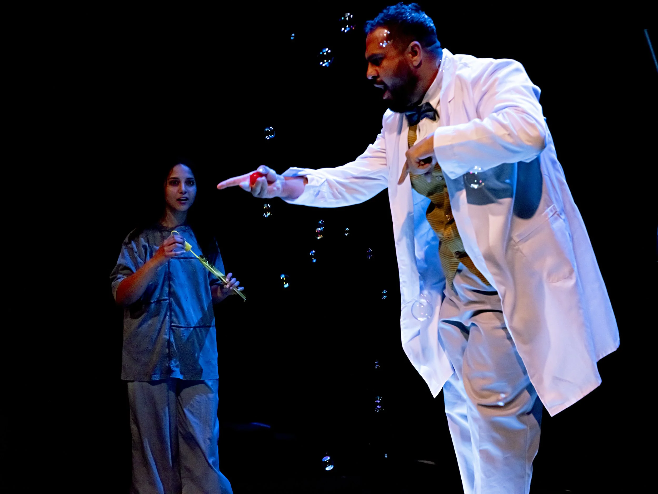 A man dressed as a scientist or magician in a white lab coat, bow tie, and yellow shirt is playfully pointing at a woman with a bubble wand, creating bubbles in a dark setting.