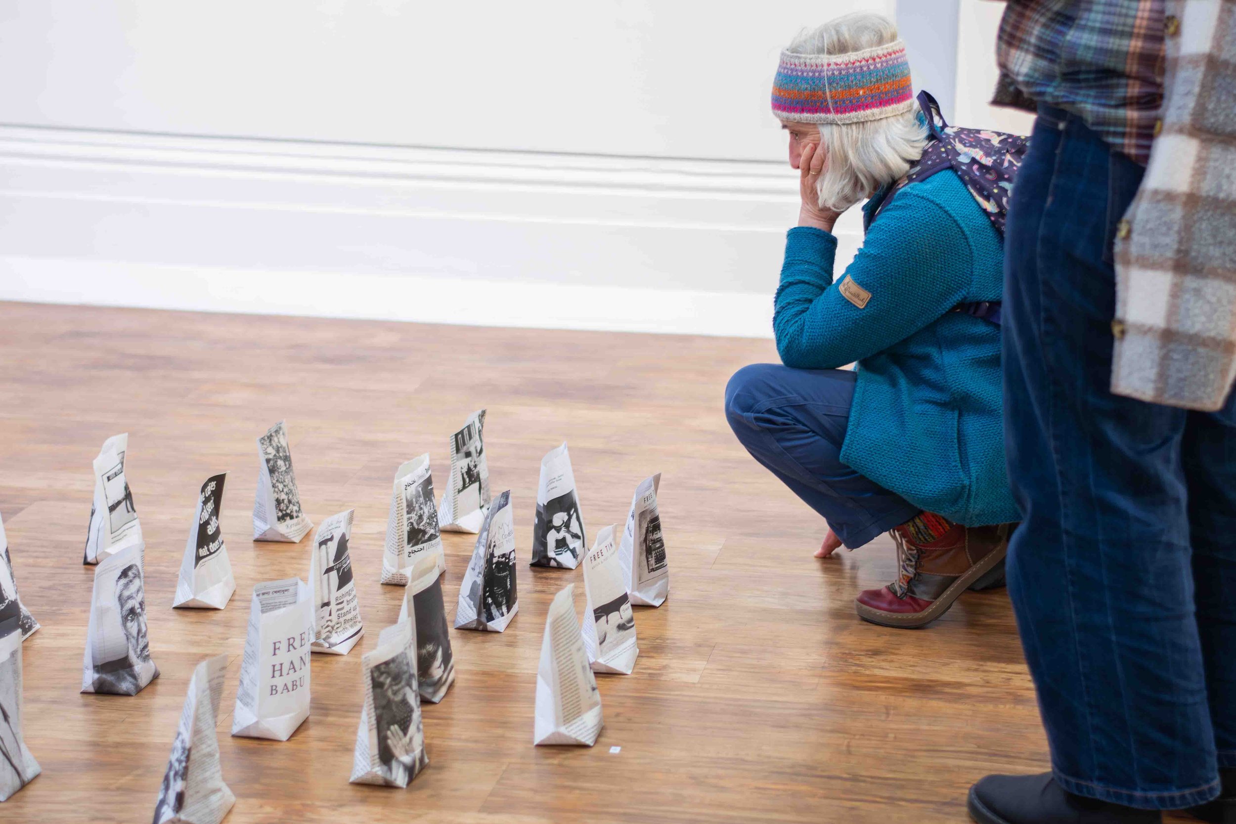 An elderly woman with gray hair, wearing a colorful knitted hat and blue jacket, kneels on the floor, looking with concern or sorrow at paper bags arranged in a row, while a person in a plaid shirt and jeans stands nearby.