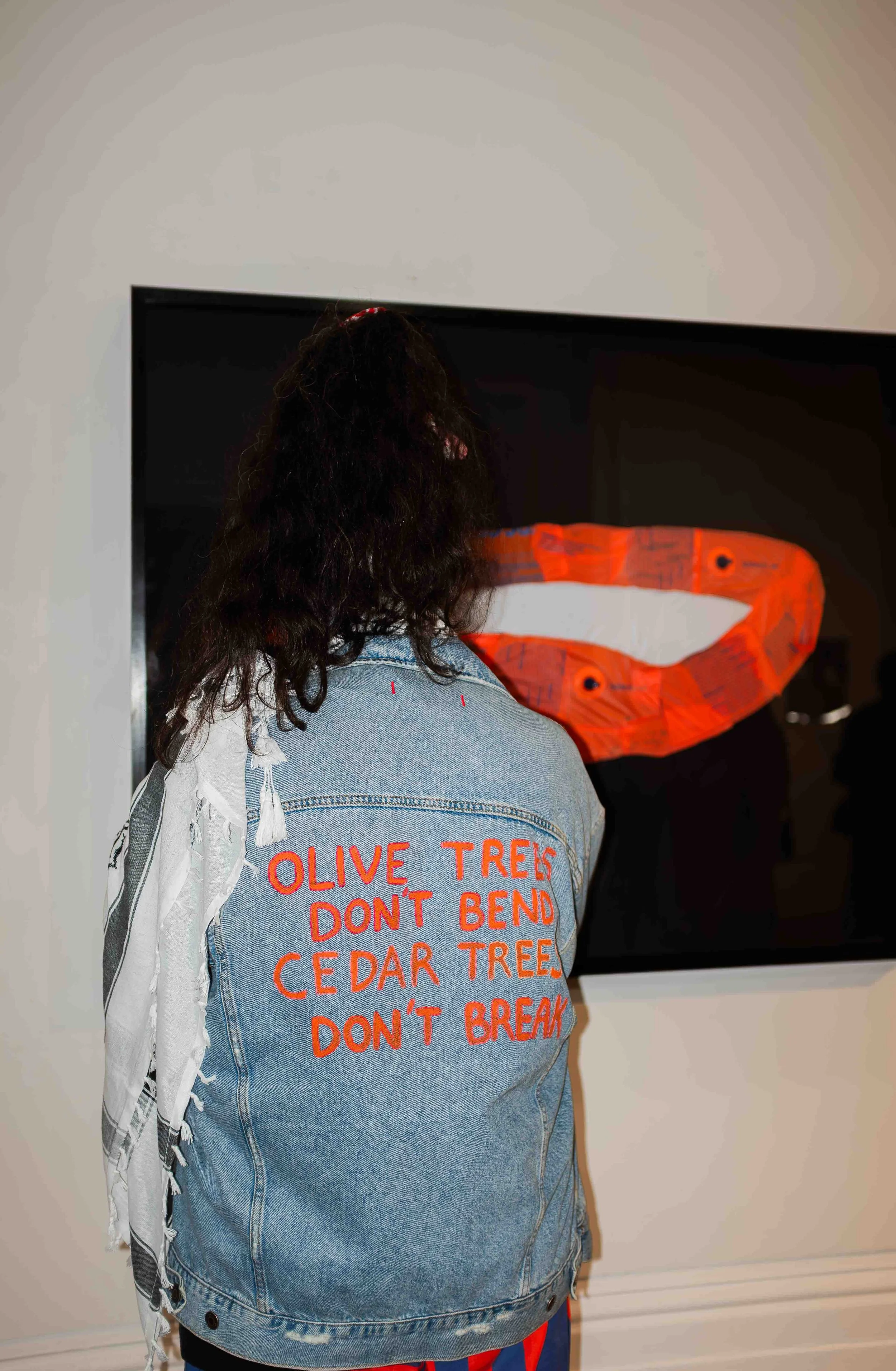 Person with long curly dark hair, wearing a denim jacket with orange text on the back, standing in front of a screen with a heart-shaped object.