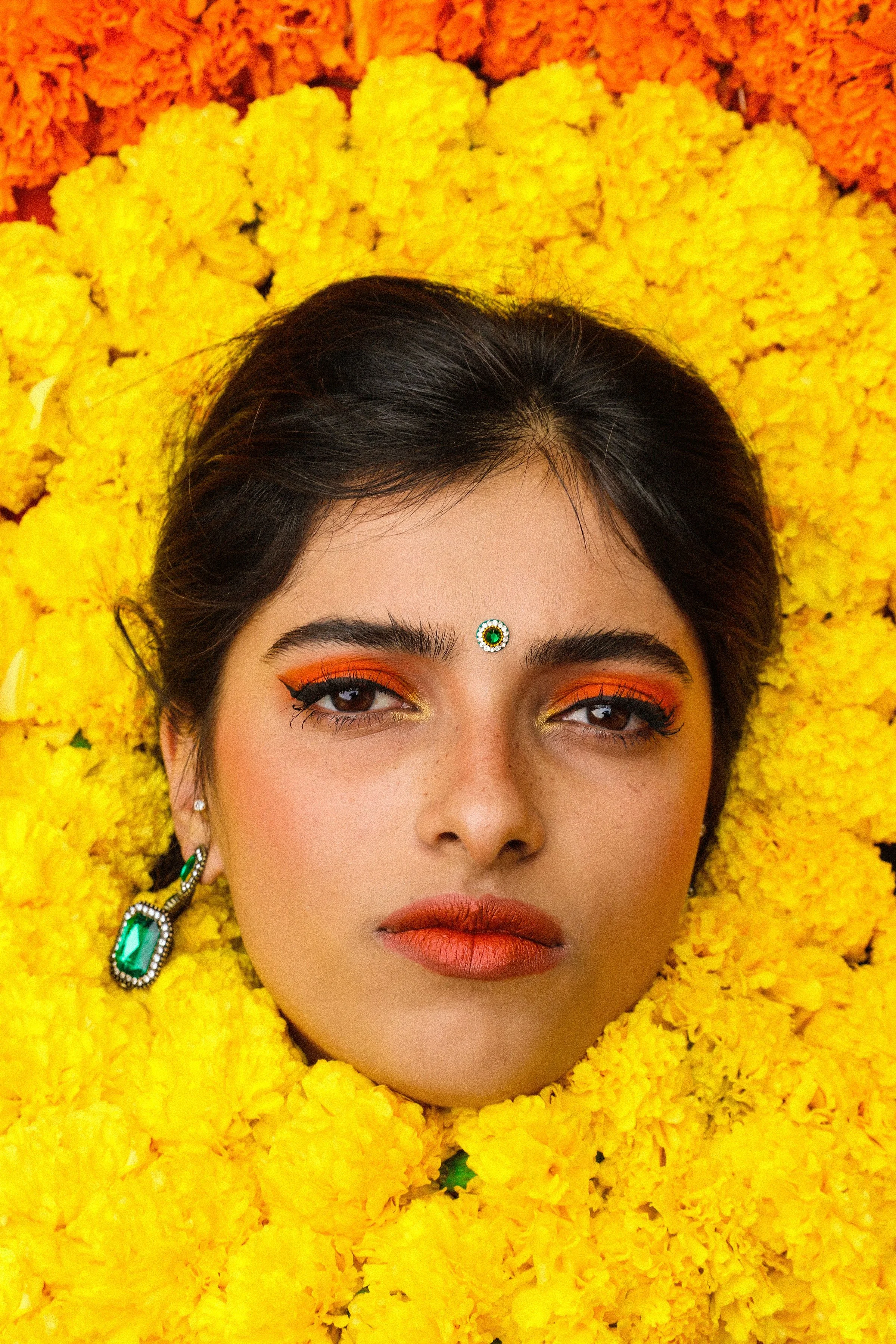 Close-up of a woman's face with makeup, jewelry, and traditional adornments, surrounded by yellow and orange marigold flowers.