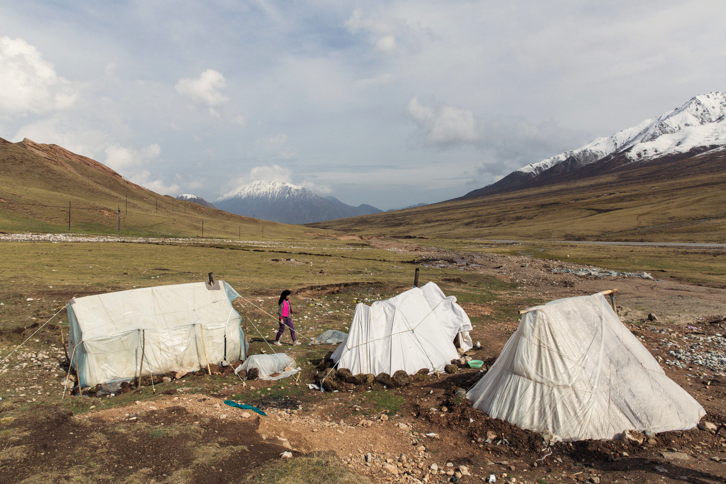 A landscape with snow-capped mountains in the background, grassy hills, and tents in the foreground with a person walking nearby.