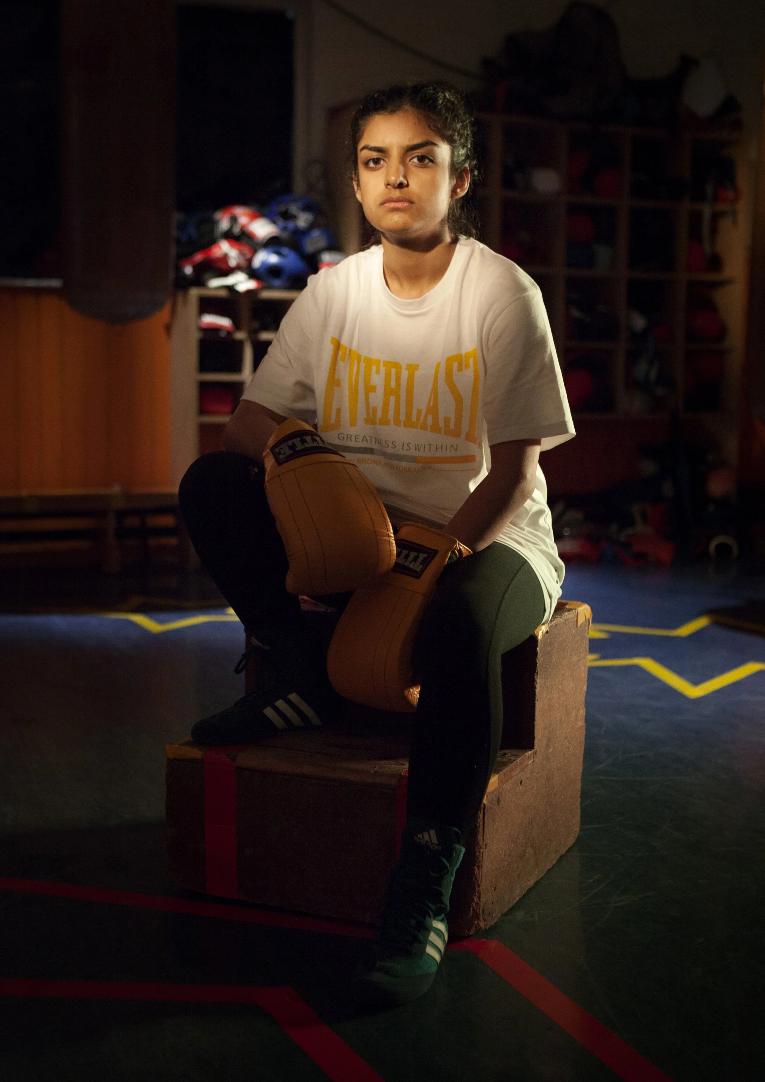 A young woman seated on a wooden box in a boxing gym, wearing a white t-shirt with yellow lettering, black leggings, and green and black sneakers, holding an orange boxing glove, with a background of boxing gloves and equipment on shelving, dimly lit