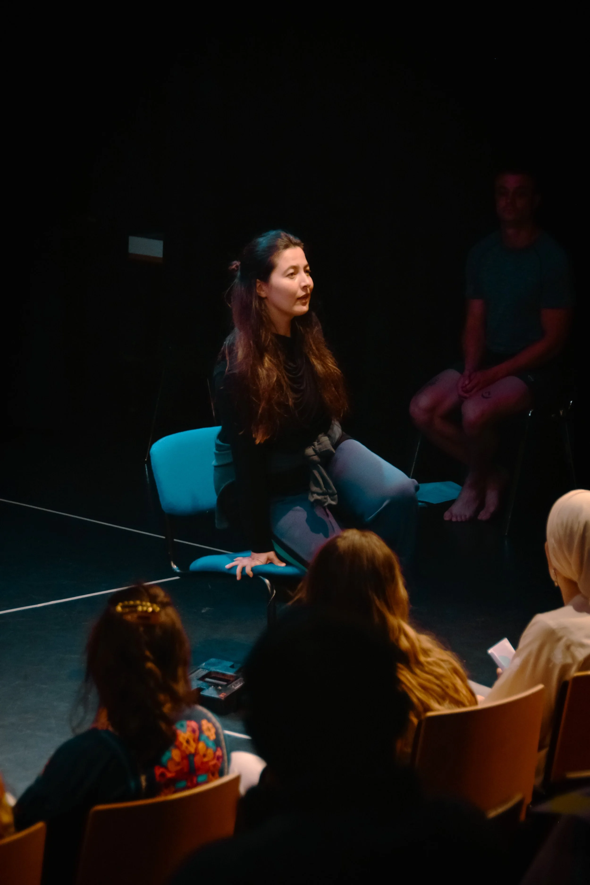 A woman with long curly hair is sitting on a chair in a dark room, speaking or performing, while a man in shorts sits nearby on a high stool or chair. Audience members are seated in front of her, and the scene appears to be in a performance or rehear
