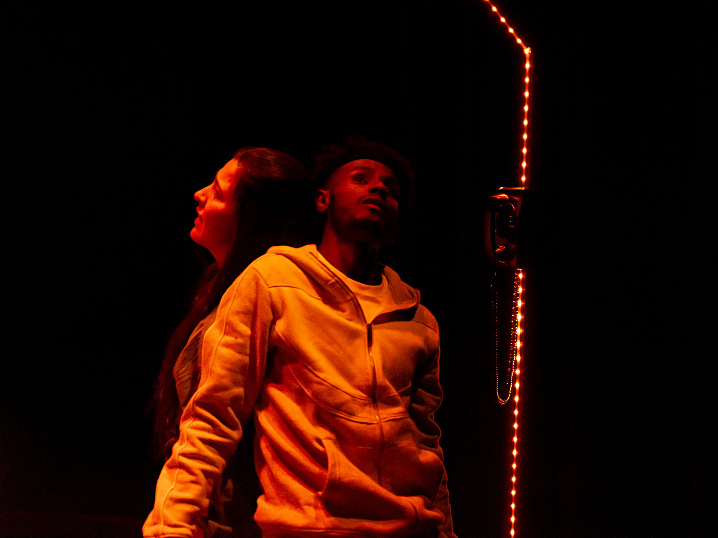 Three people stand in front of a dark background with orange lights. One woman faces left, another person with glasses faces right, and a man with an afro looks upward.
