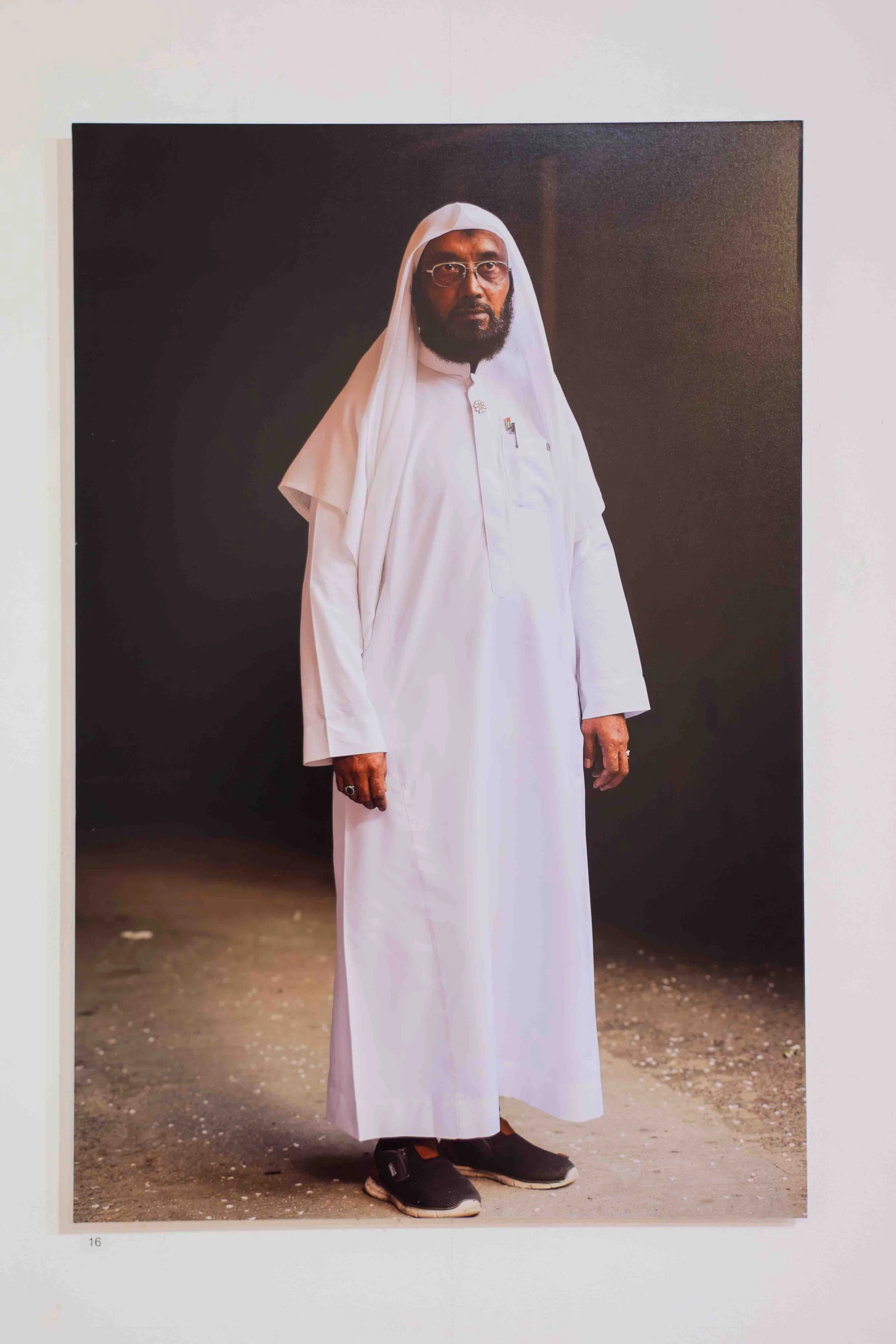 A man with dark skin, glasses, and a full beard wearing traditional white Middle Eastern attire standing outdoors on a dirt ground, with a black background and a white wall behind him.