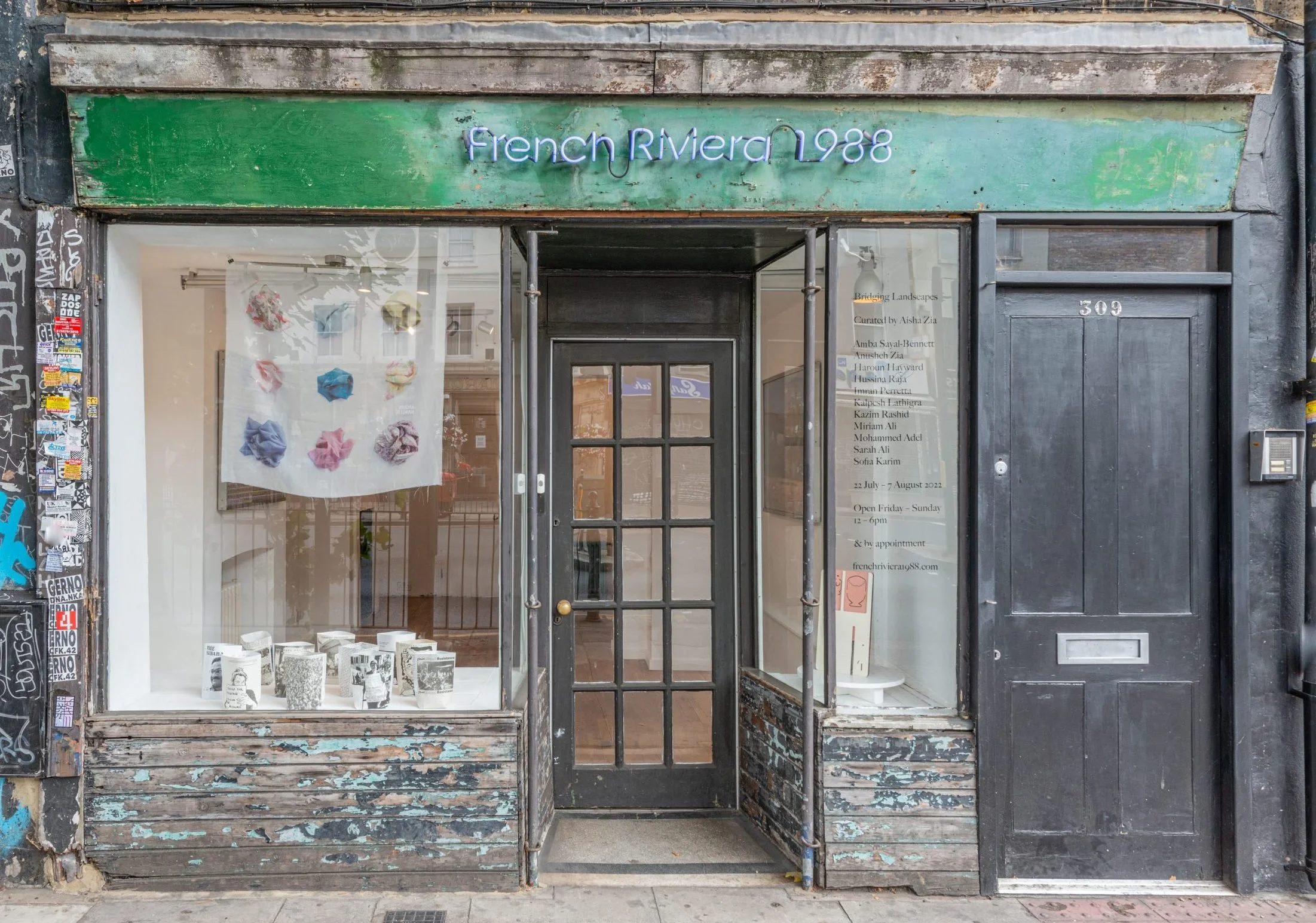 storefront with a green sign that reads 'french RIVERA 1988' in purple neon, with a window display of various black and white books or magazines, and advertising or informational text on the right side of the entrance.