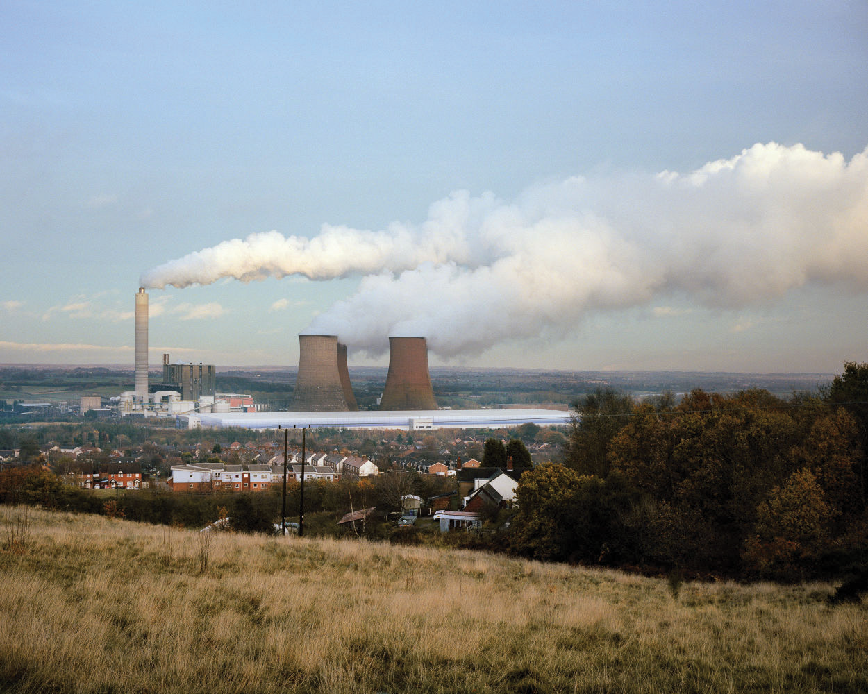 A landscape view of a power plant with cooling towers and smokestacks releasing smoke, surrounded by residential houses and greenery.
