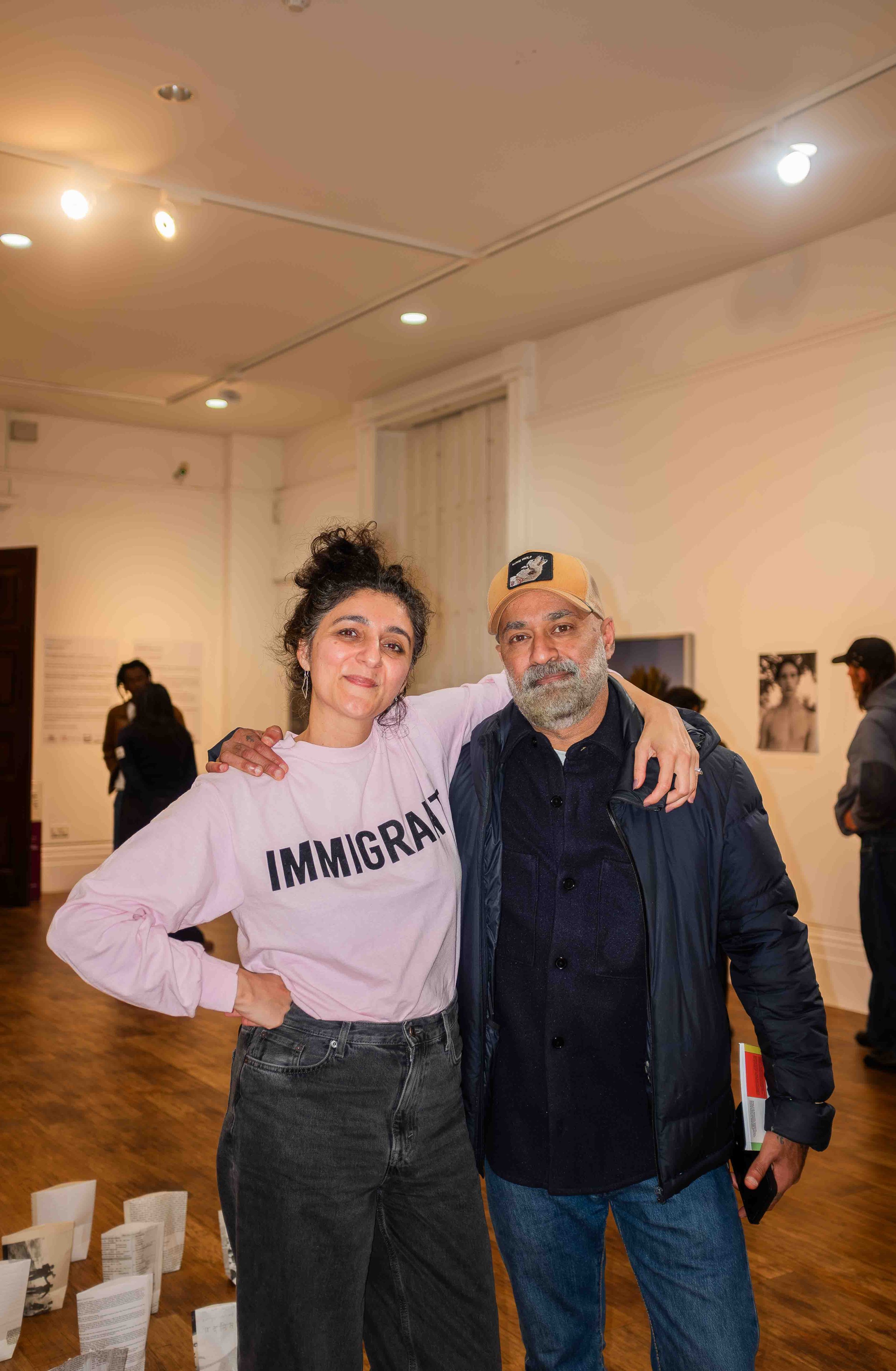 A woman wearing a white shirt with the word 'IMMIGRANT' and a man wearing a beige cap and black jacket smiling and standing close together in an indoor event space with other people in the background.