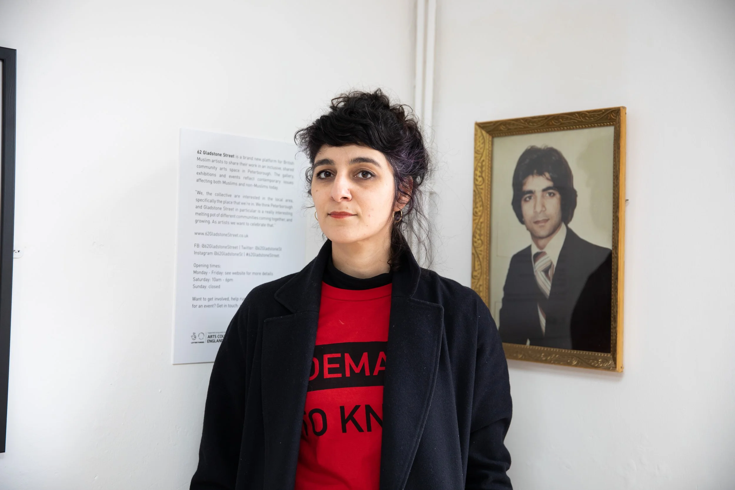 A woman with dark, curly hair and light skin standing indoors in front of a white wall. Behind her, there is a framed portrait of a man in a suit and tie, and a poster with text about a community arts space.