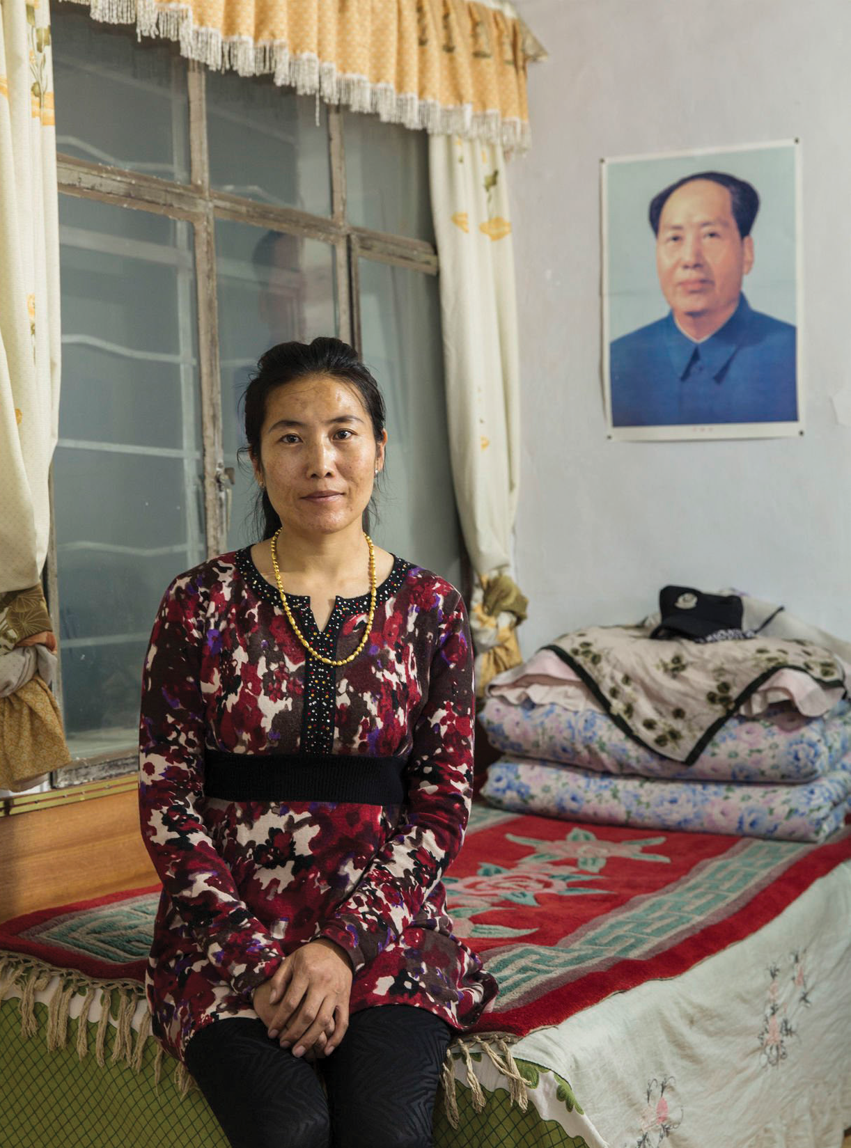 A woman sitting on a bed in a room with a portrait of Mao Zedong on the wall. The woman is wearing a floral dress with a black belt and gold necklace. Behind her are pillows and blankets on the bed, and the room has curtains and a window.