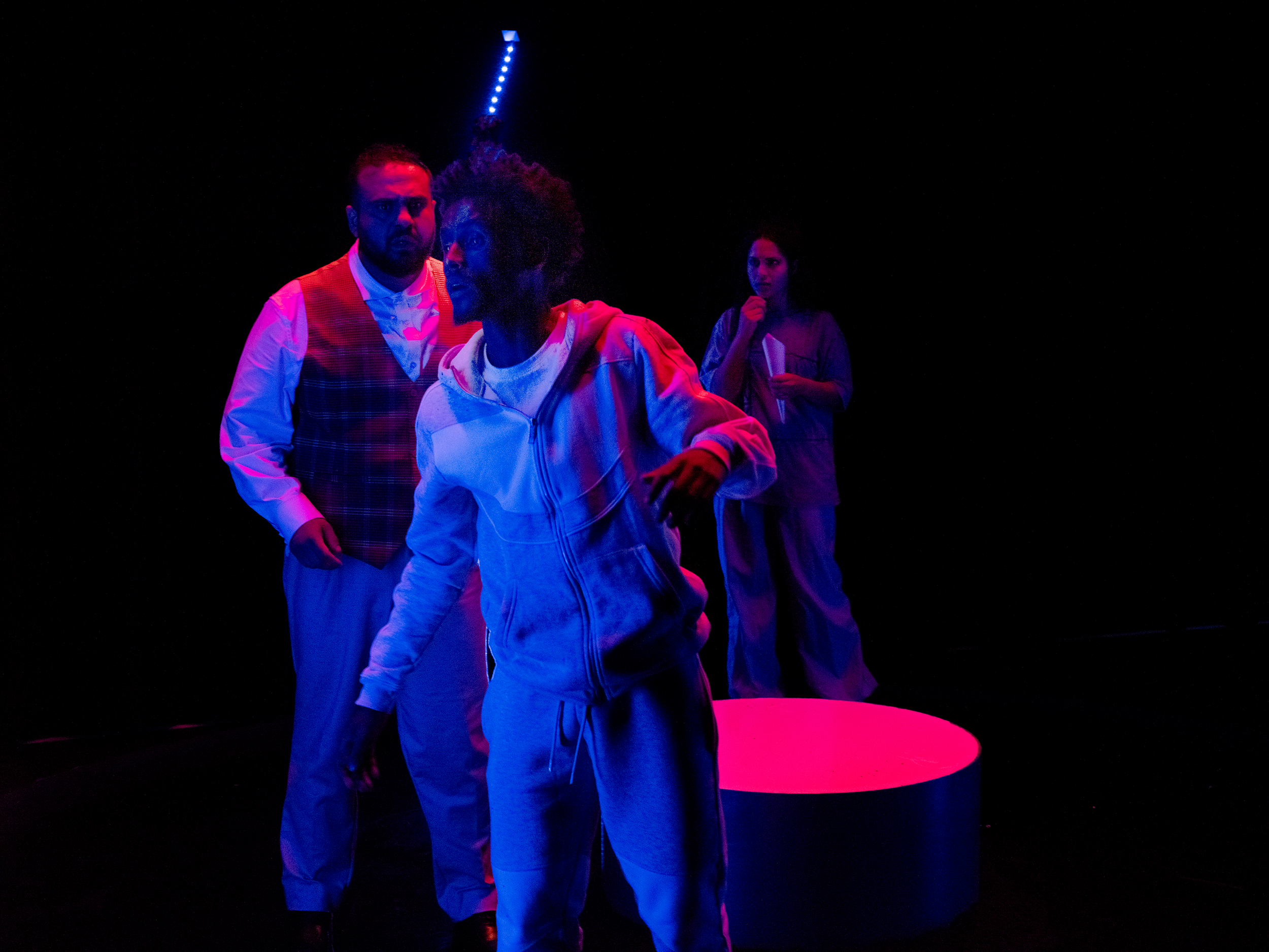 Three people standing in a dark room illuminated by colored lights. The man on the left wears a vest and has facial hair, looking serious. The woman in the center is wearing a hoodie, with an afro hairstyle, looking to her left. The woman on the righ