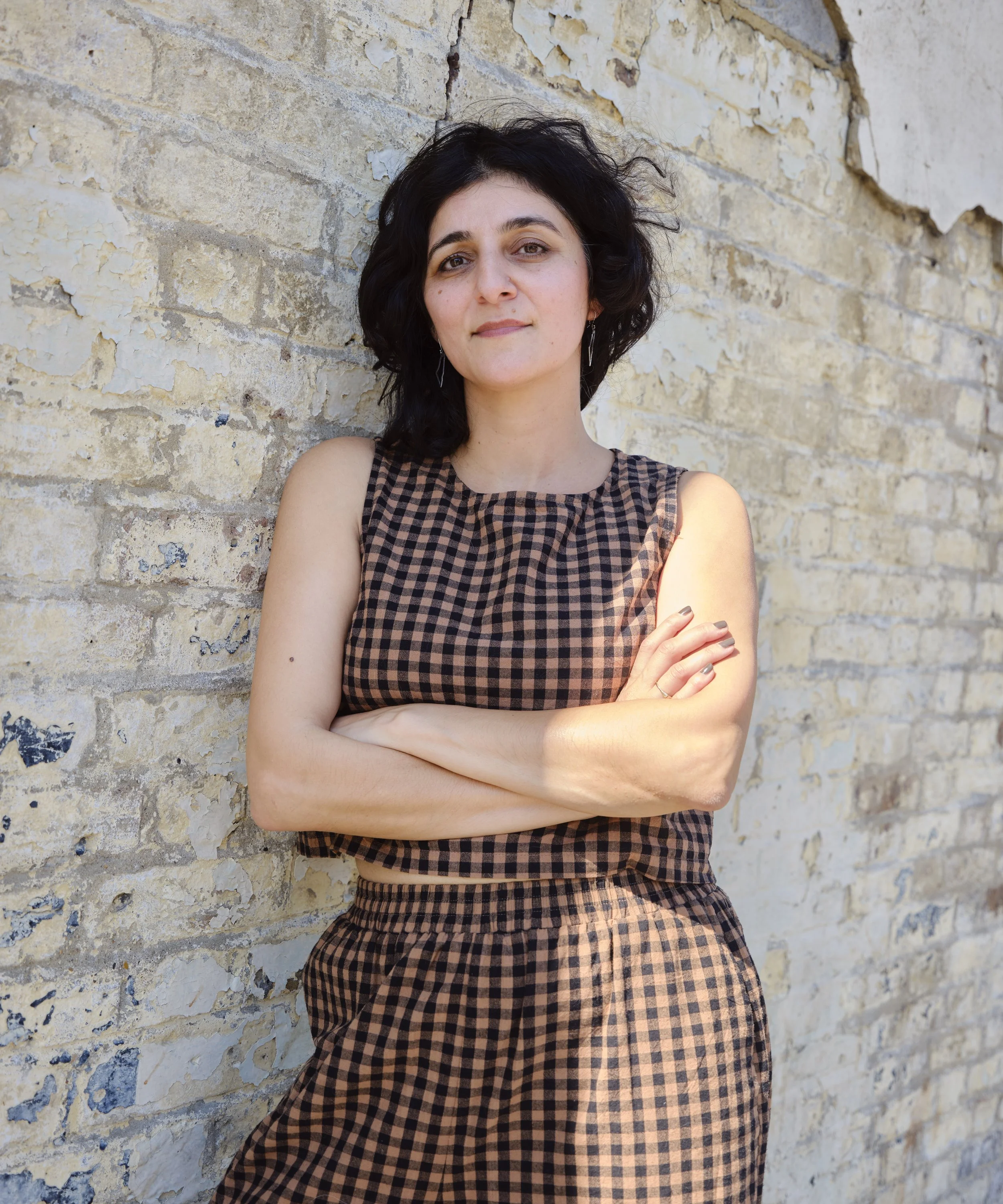 A woman with short black curly hair posing with crossed arms against a weathered brick wall.
