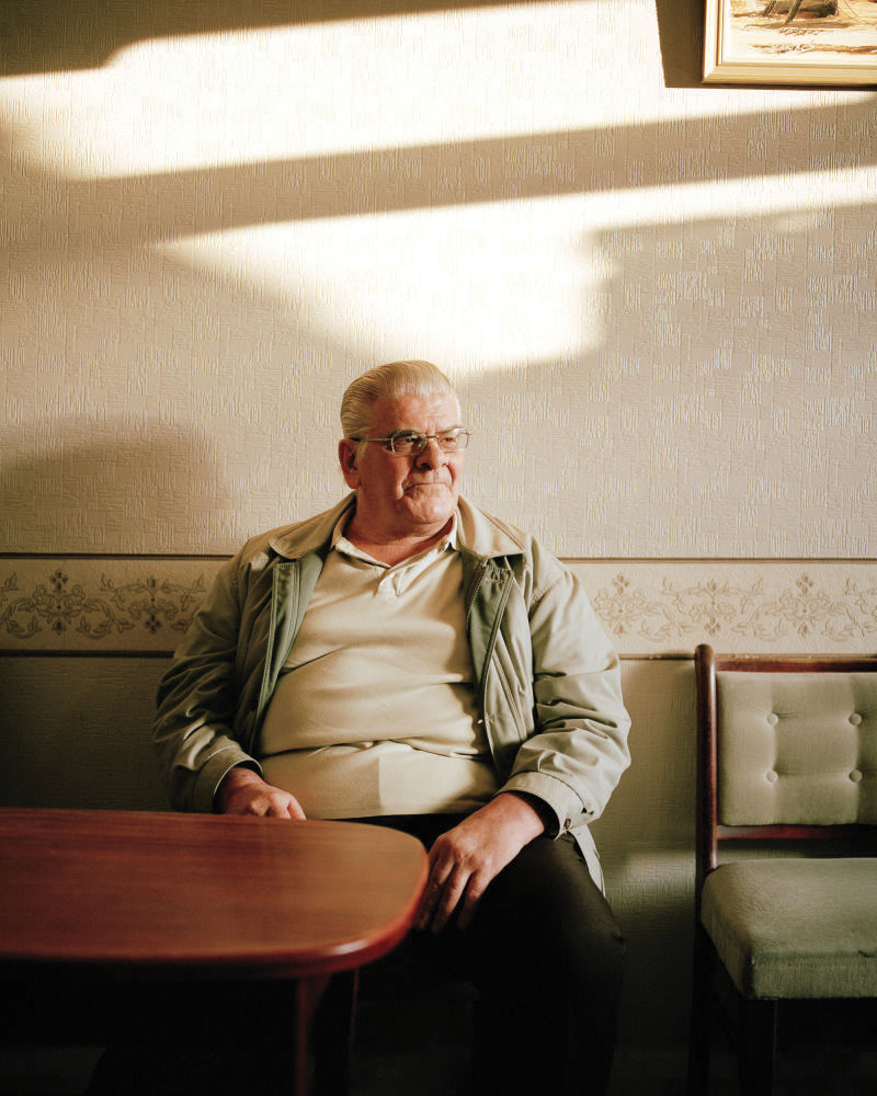 Older man with glasses and slicked-back gray hair sitting alone in a booth at a restaurant or cafe, lit by sunlight coming through a window, with a painting partially visible on the wall.
