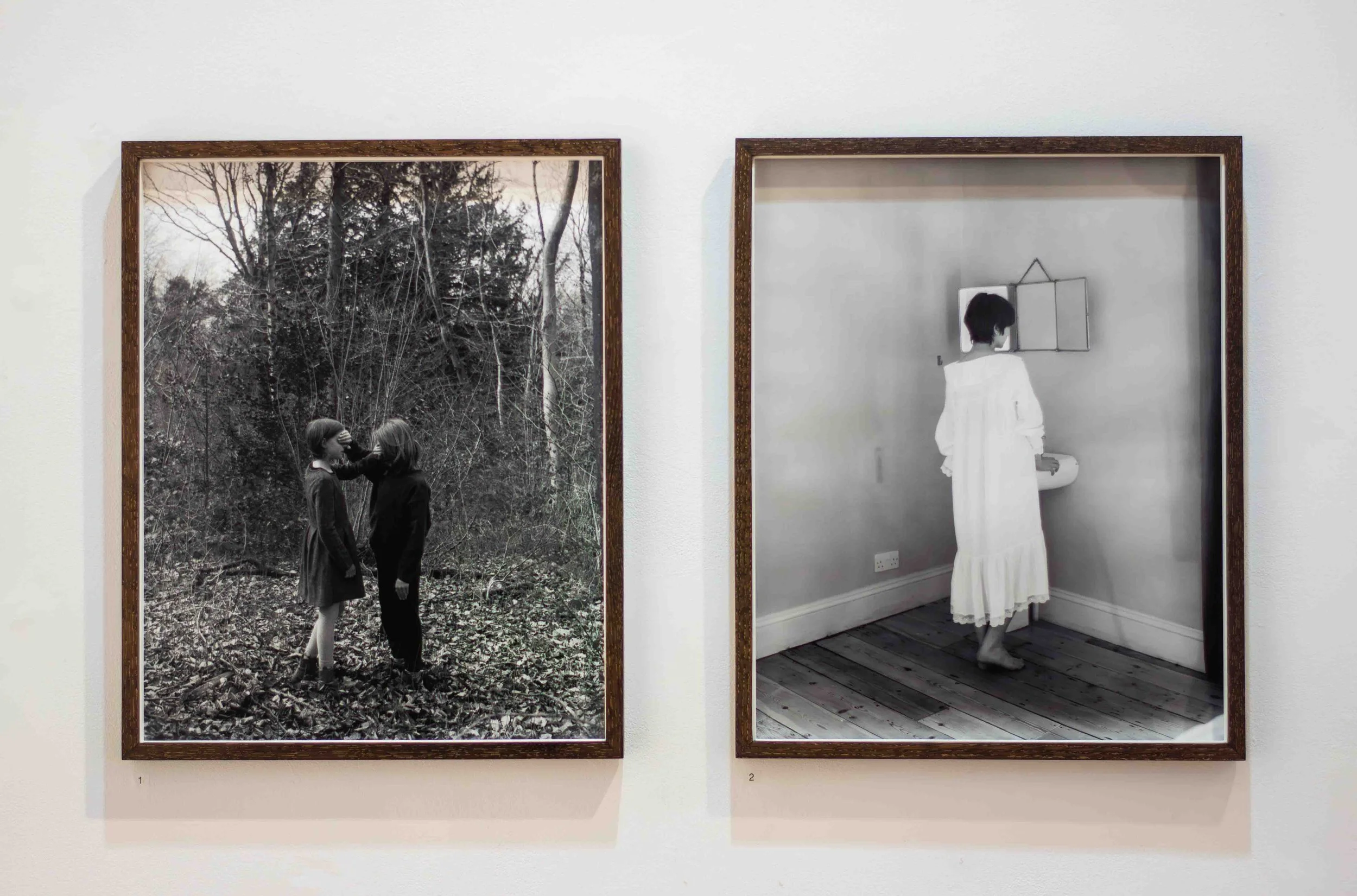 Two framed black-and-white photographs hanging on a white wall. The left photo depicts two children, one with dark hair and one with light hair, playing among trees in a forest. The right photo shows a woman in a long white dress standing inside a ro