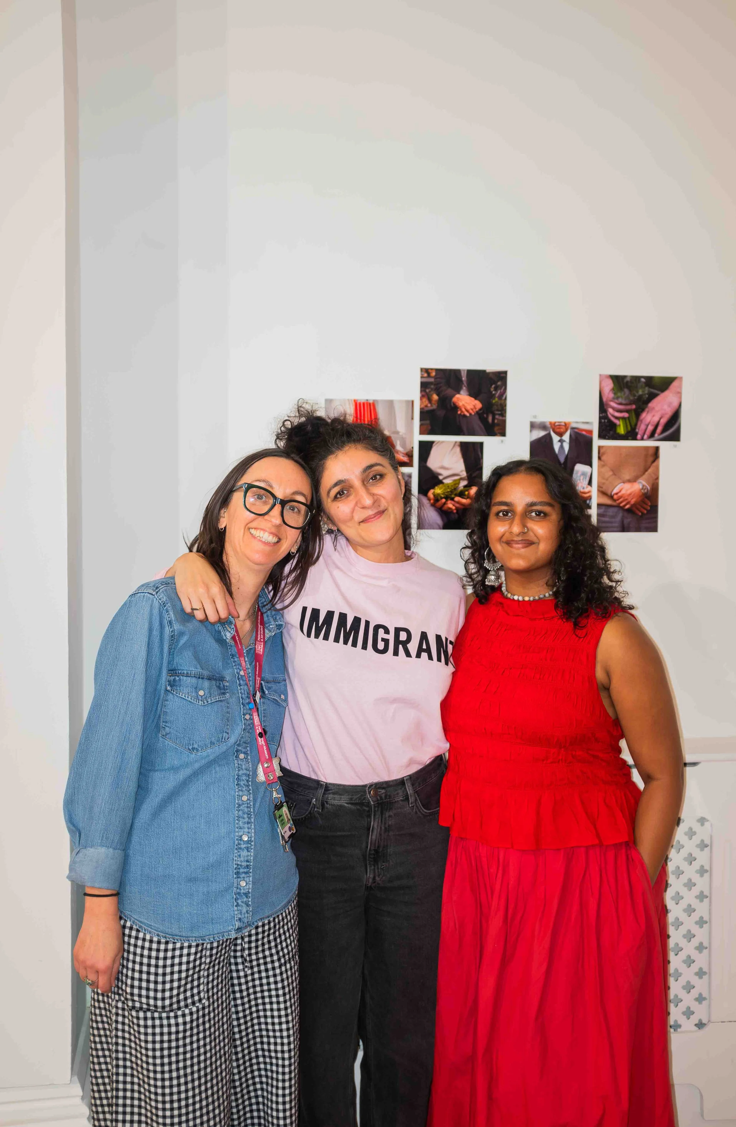 Three women standing together, smiling, with photos on the wall behind them. The woman on the left wears glasses and a denim jacket, the woman in the middle wears a pink shirt with the word 'IMMIGRANT' on it, and the woman on the right wears a red dr