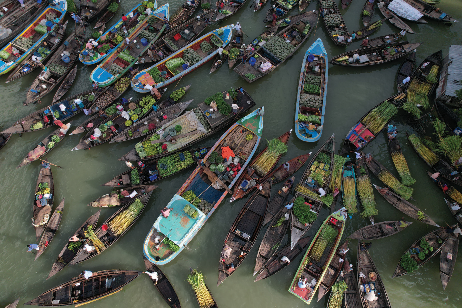 Overhead view of numerous small boats in a floating market, each filled with various fruits, vegetables, and goods, with vendors and shoppers moving among them.