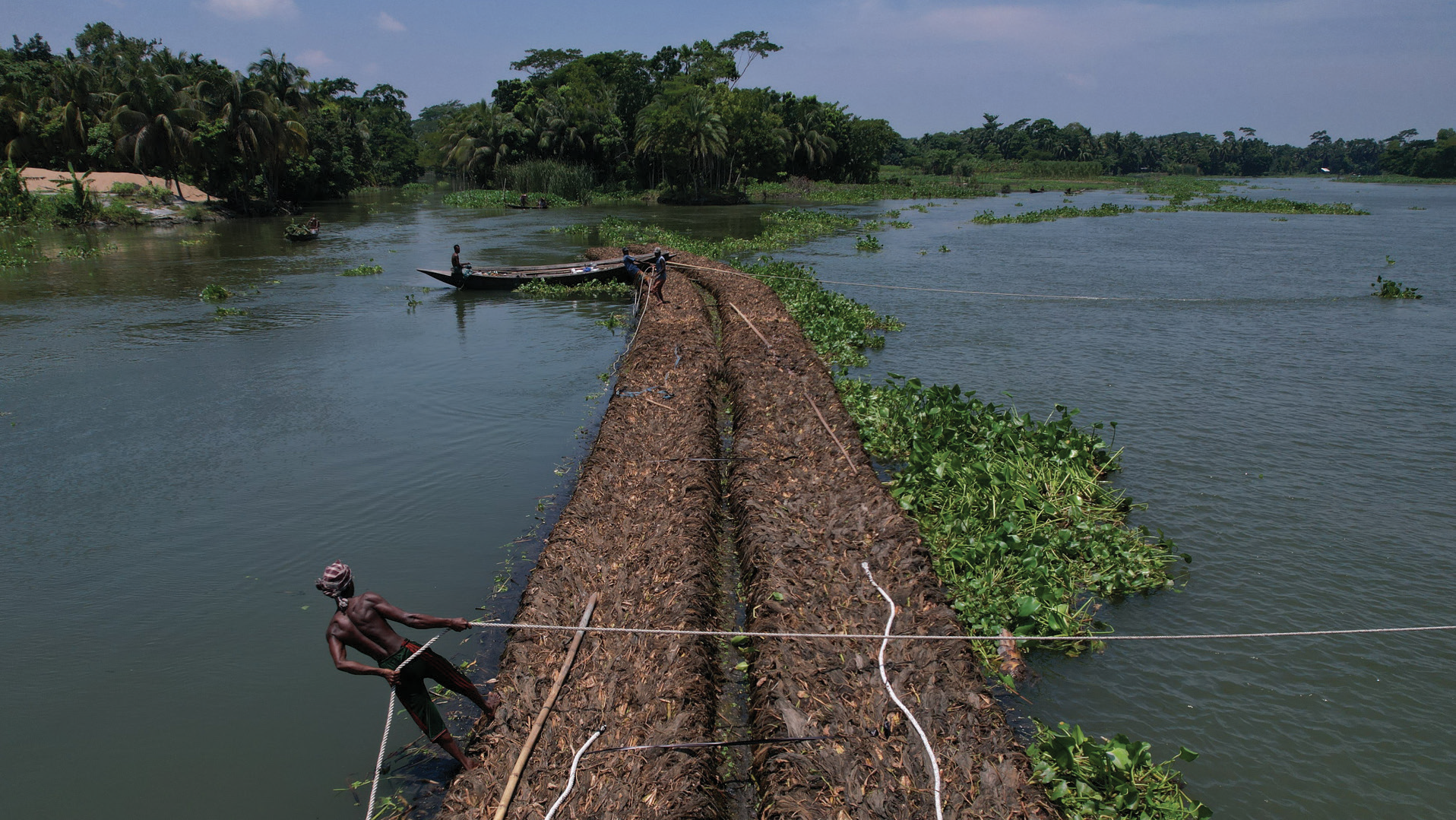 Men laying logs across a river to create a floating bridge, with boats and lush green trees in the background.