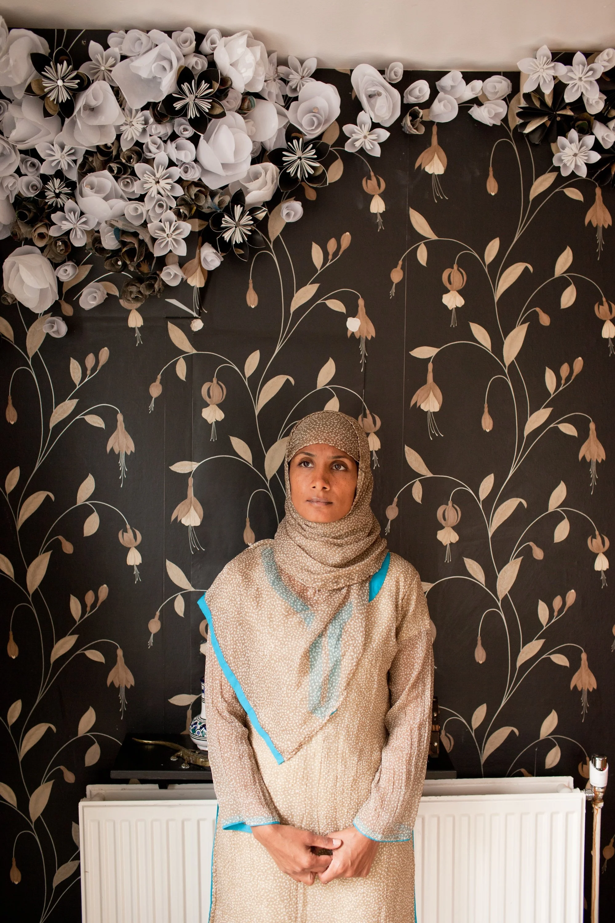 A woman wearing a headscarf and a traditional dress standing in front of a black wall with floral patterns and paper flowers.