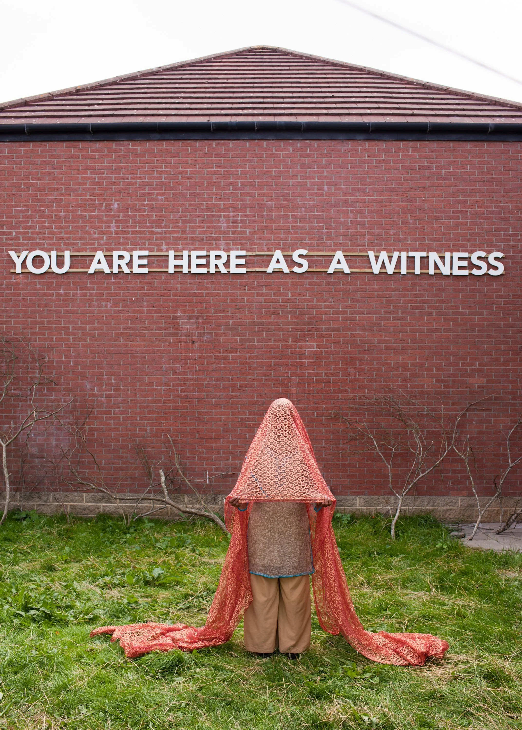 Person standing in front of a brick wall with white letters that say 'You are here as a witness.' The person is holding a large, red patterned fabric that covers their head and shoulders, and they are wearing beige pants and a grey and turquoise top.