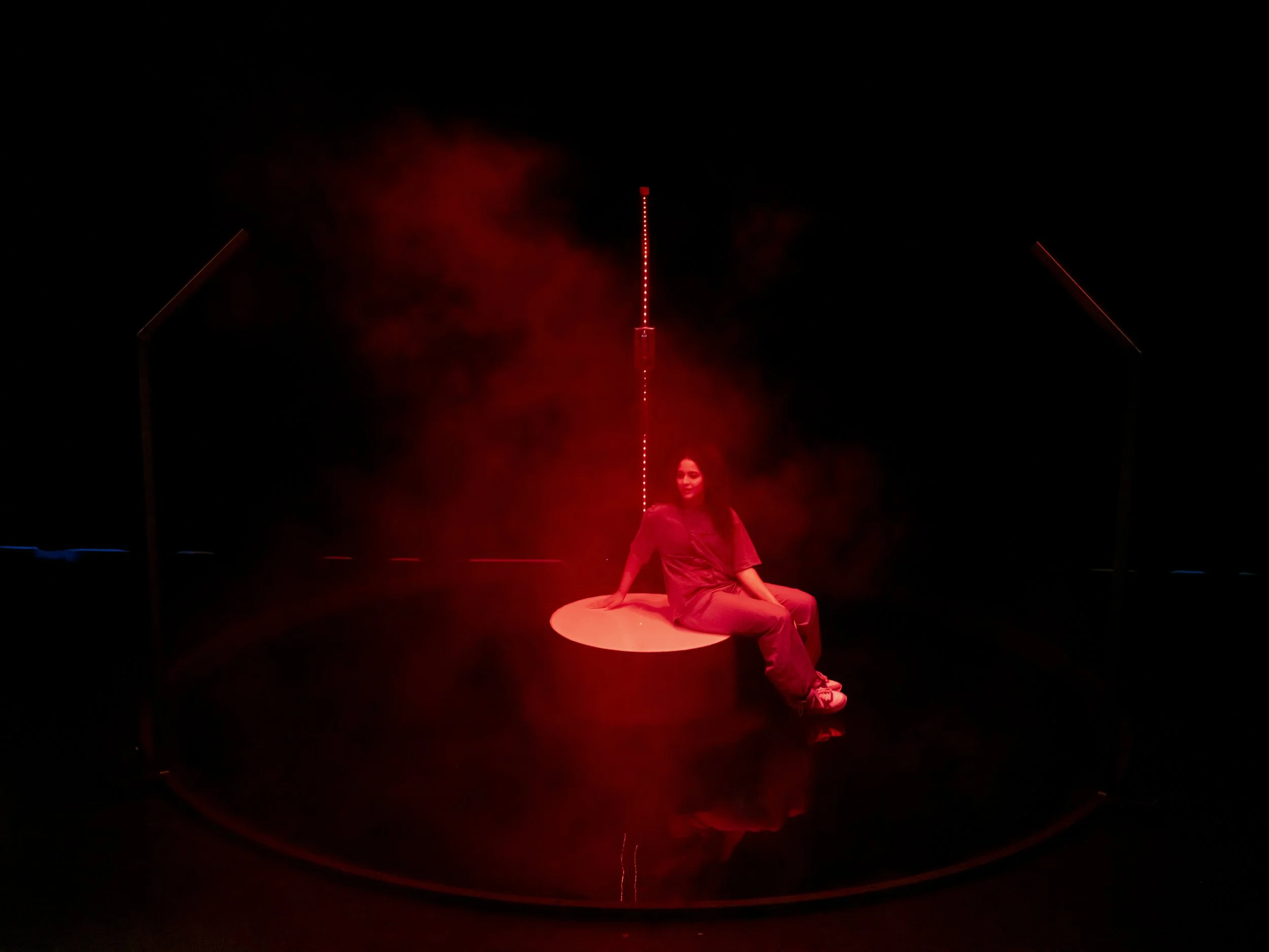 A female performer sitting on a circular platform with red lighting and smoke effects, surrounded by vertical red laser beams in a dark setting.