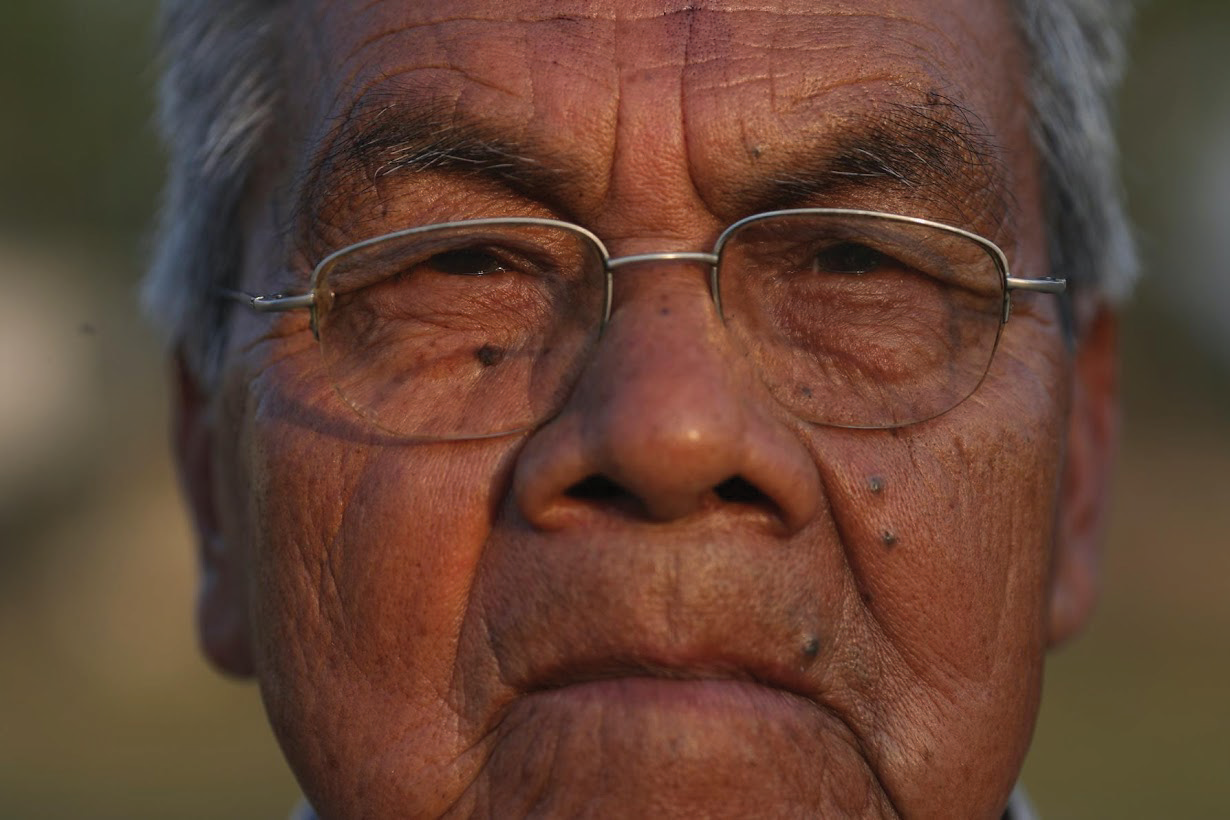 Close-up of an elderly man's face with tanned skin, glasses, gray hair, and visible wrinkles.