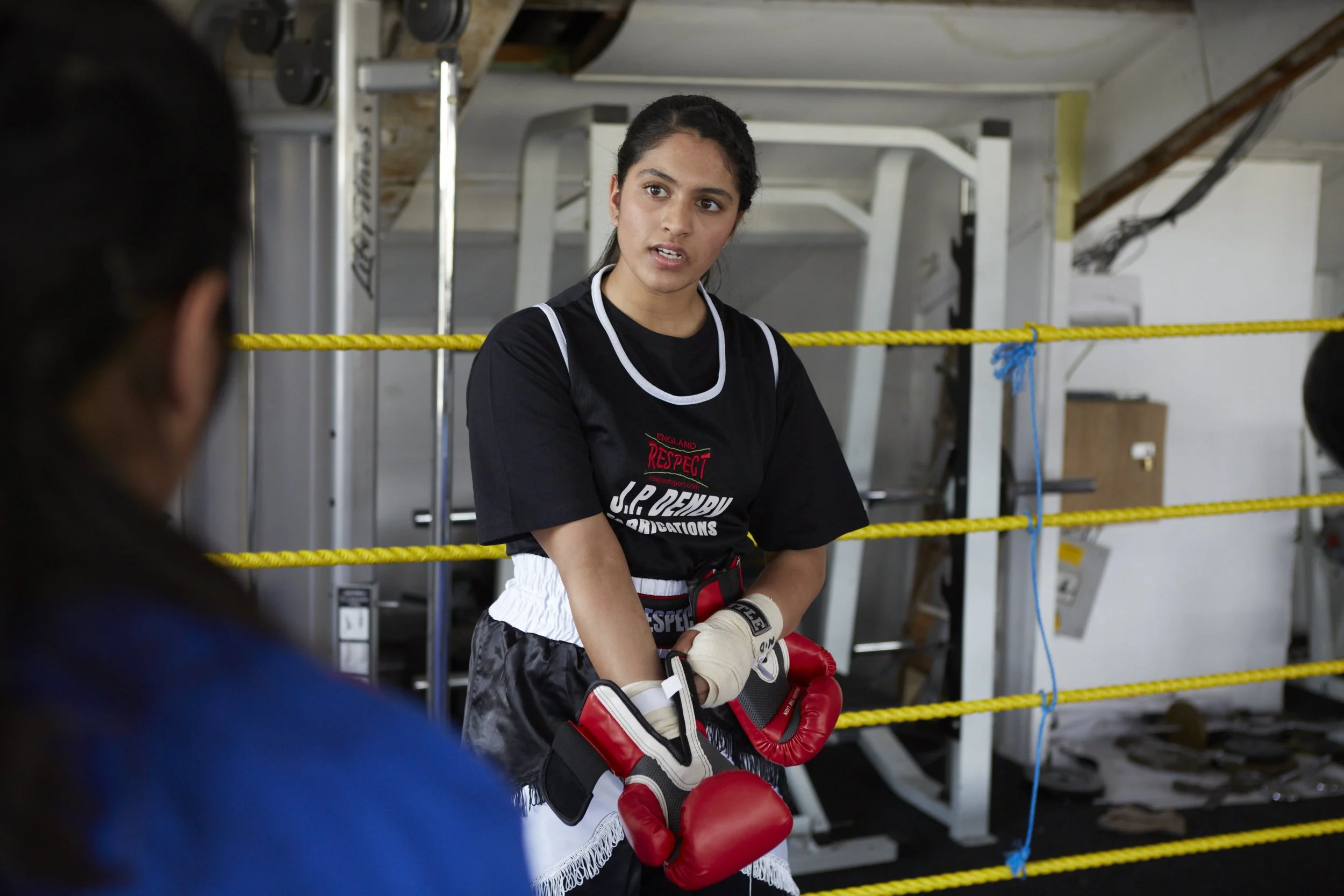 A female boxer in boxing gloves and sports attire sitting on a bench, listening attentively during a training session inside a gym with boxing equipment and yellow safety ropes.