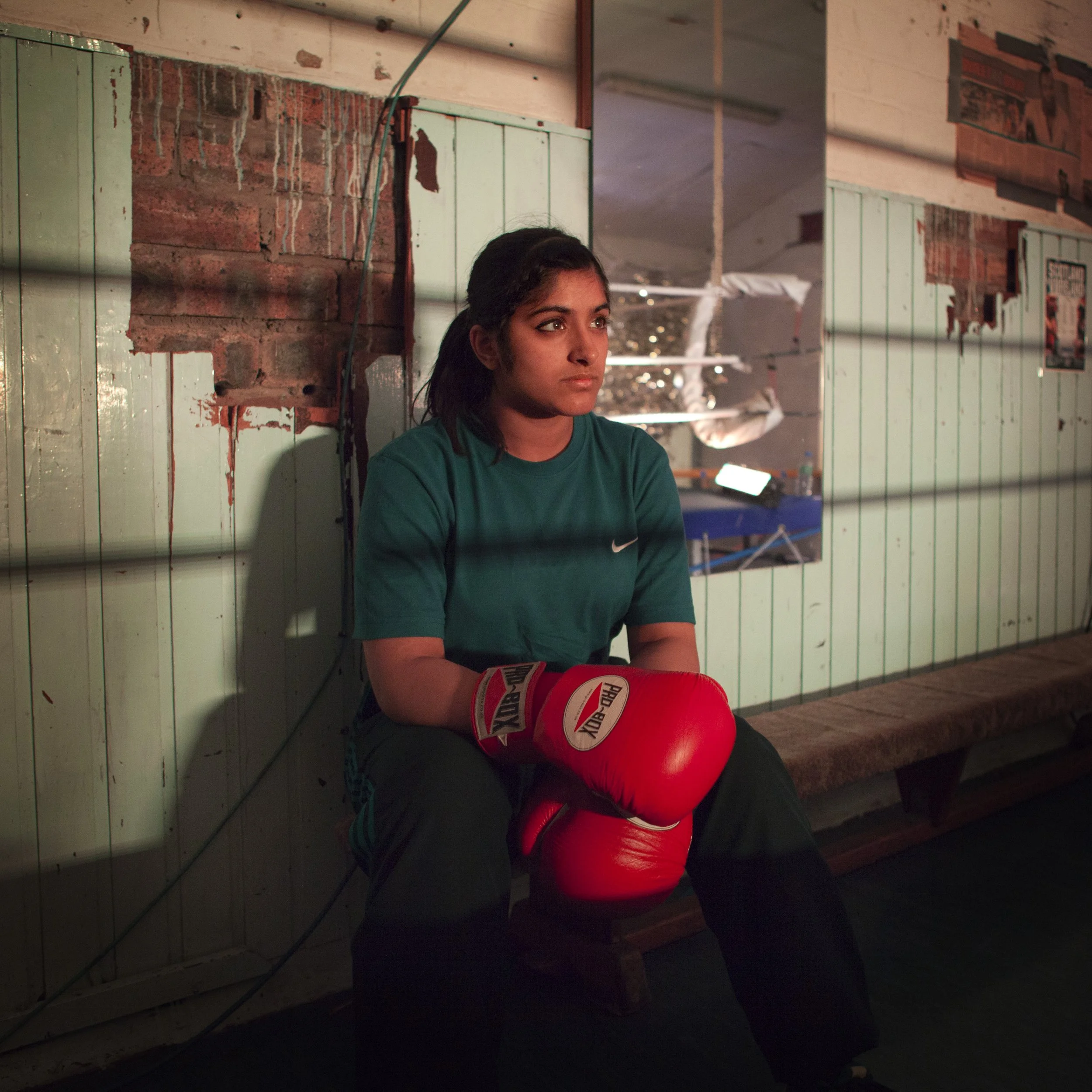 A young woman with dark hair and a teal shirt is sitting on the floor next to a red boxing glove with the brand name "Everlast." She appears to be rest or waiting with a calm, focused expression. The background features a rustic brick and wooden wall