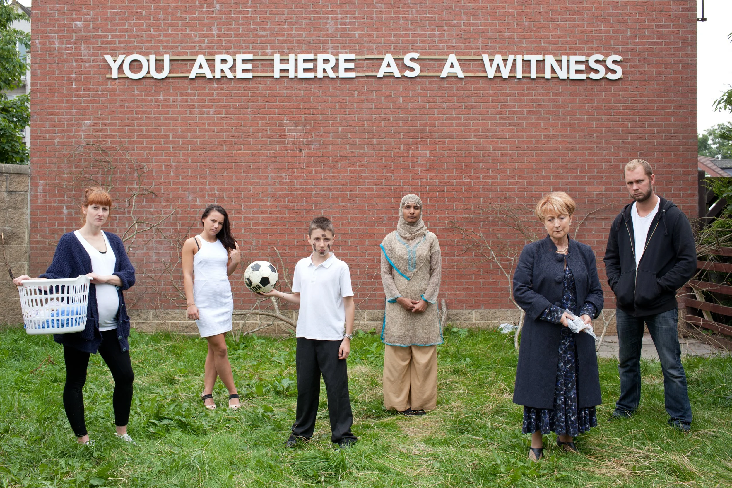 Group of seven diverse people standing outdoors in front of a brick wall with a sign that says "You Are Here As A Witness." Some people look serious or sad, and one person is holding a basket, another a soccer ball, and another a tissue.